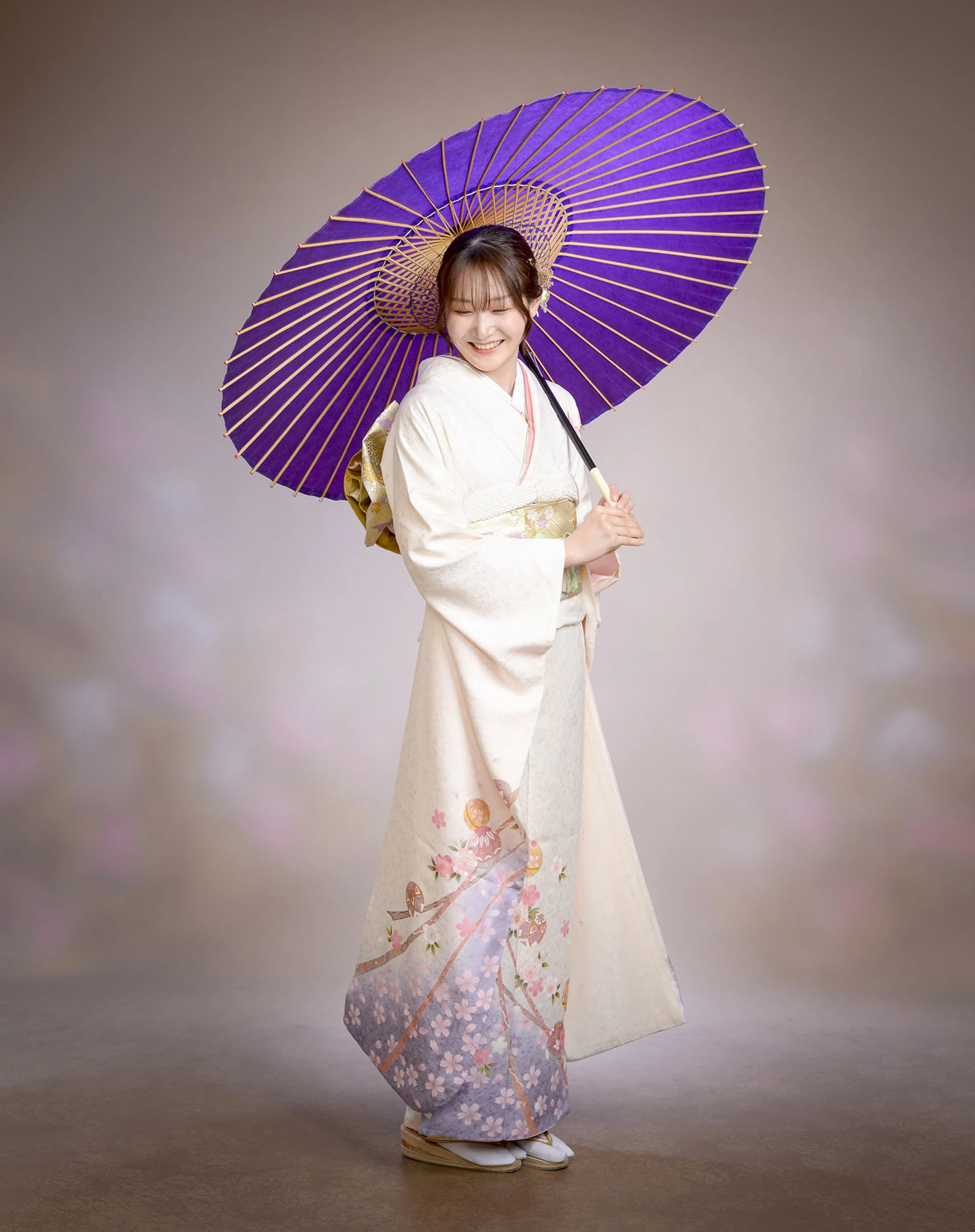 紫色の和傘をさして笑顔の若い女性。白い着物に桜と梅の花の模様。