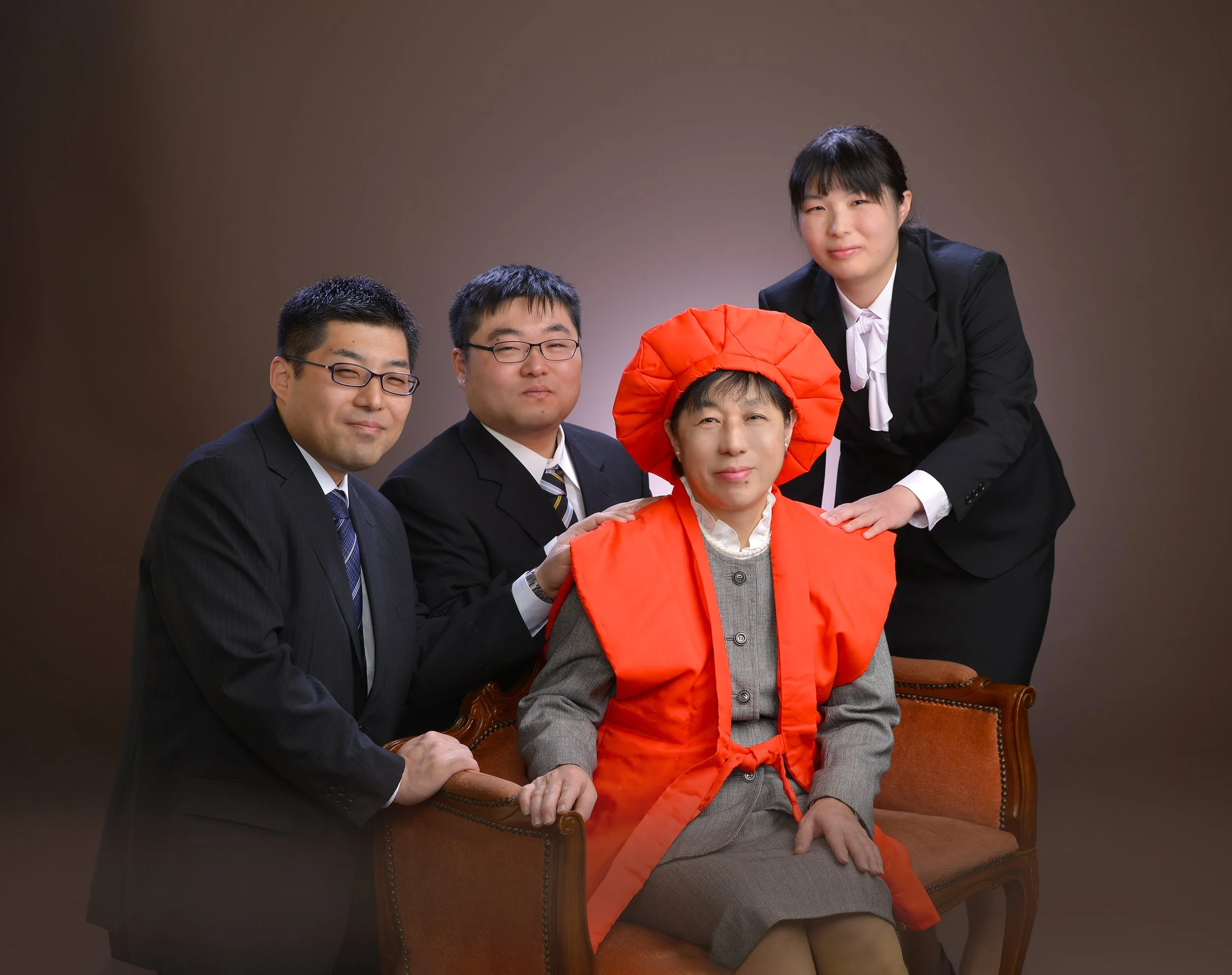 ビジネスフォーマルの服を着た4人の男女がポーズをとるグループ写真、中央の女性は赤い伝統的な衣装と帽子を着用している。