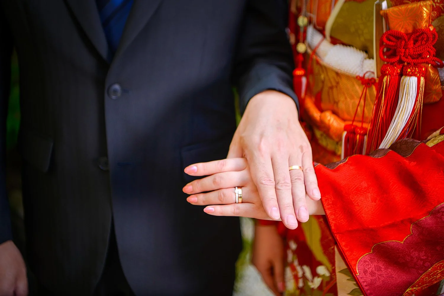 結婚式の新郎と新婦の手が重ねられており、結婚指輪が見える。新婦は伝統的な和装を着ている。