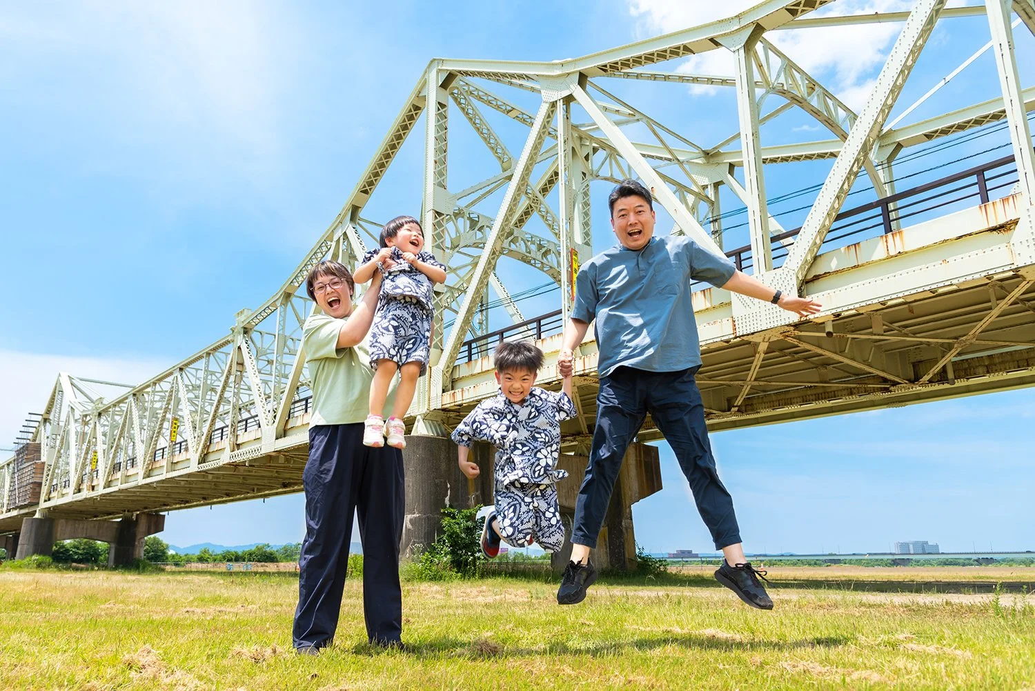 家族が橋の下の草原の上でジャンプしている写真。青い空と鉄橋が背景に広がる。
