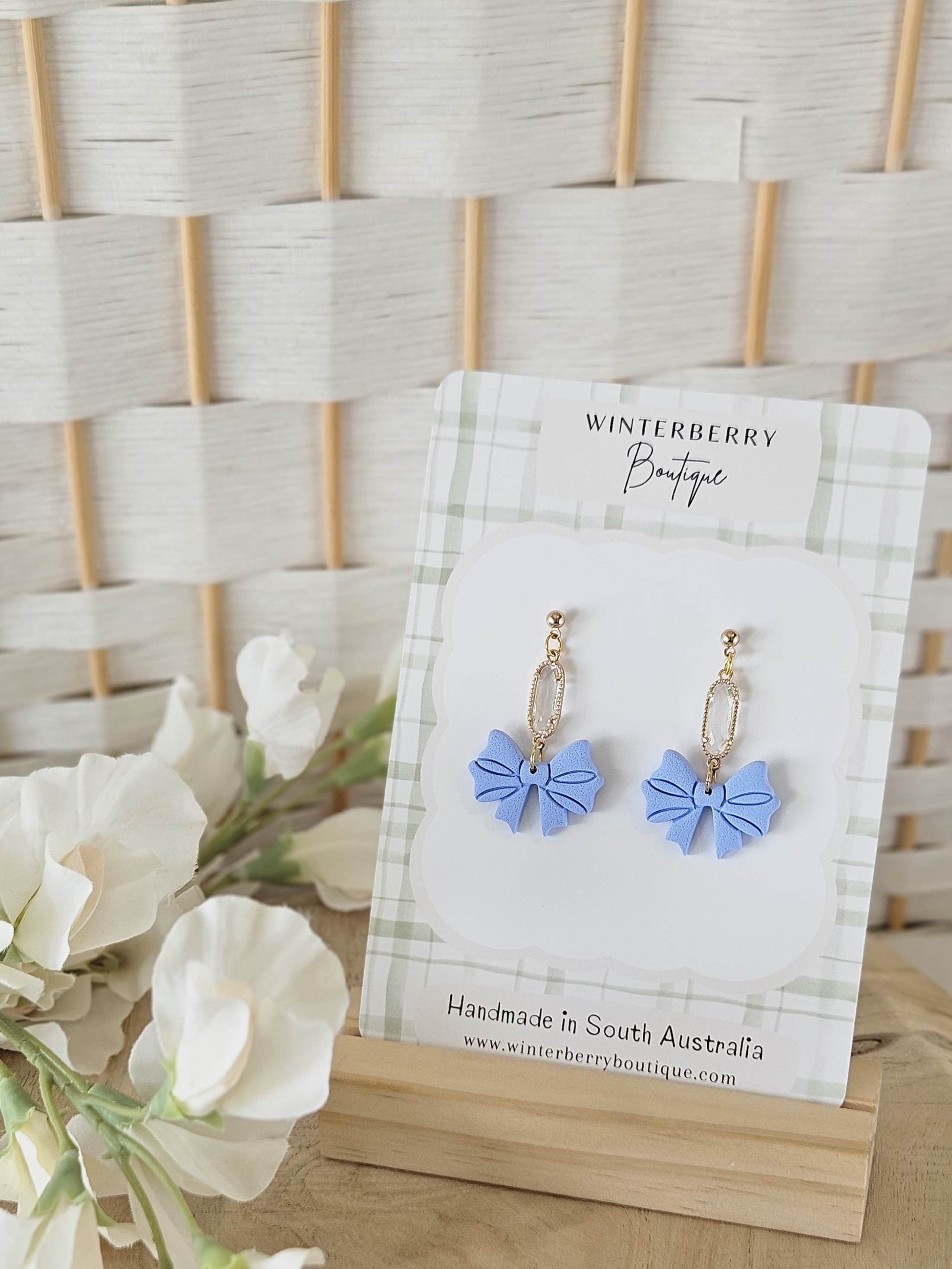 Baby blue polymer clay bow earrings with crystal accents on a plaid card displayed with white flowers and a cream lattice backdrop