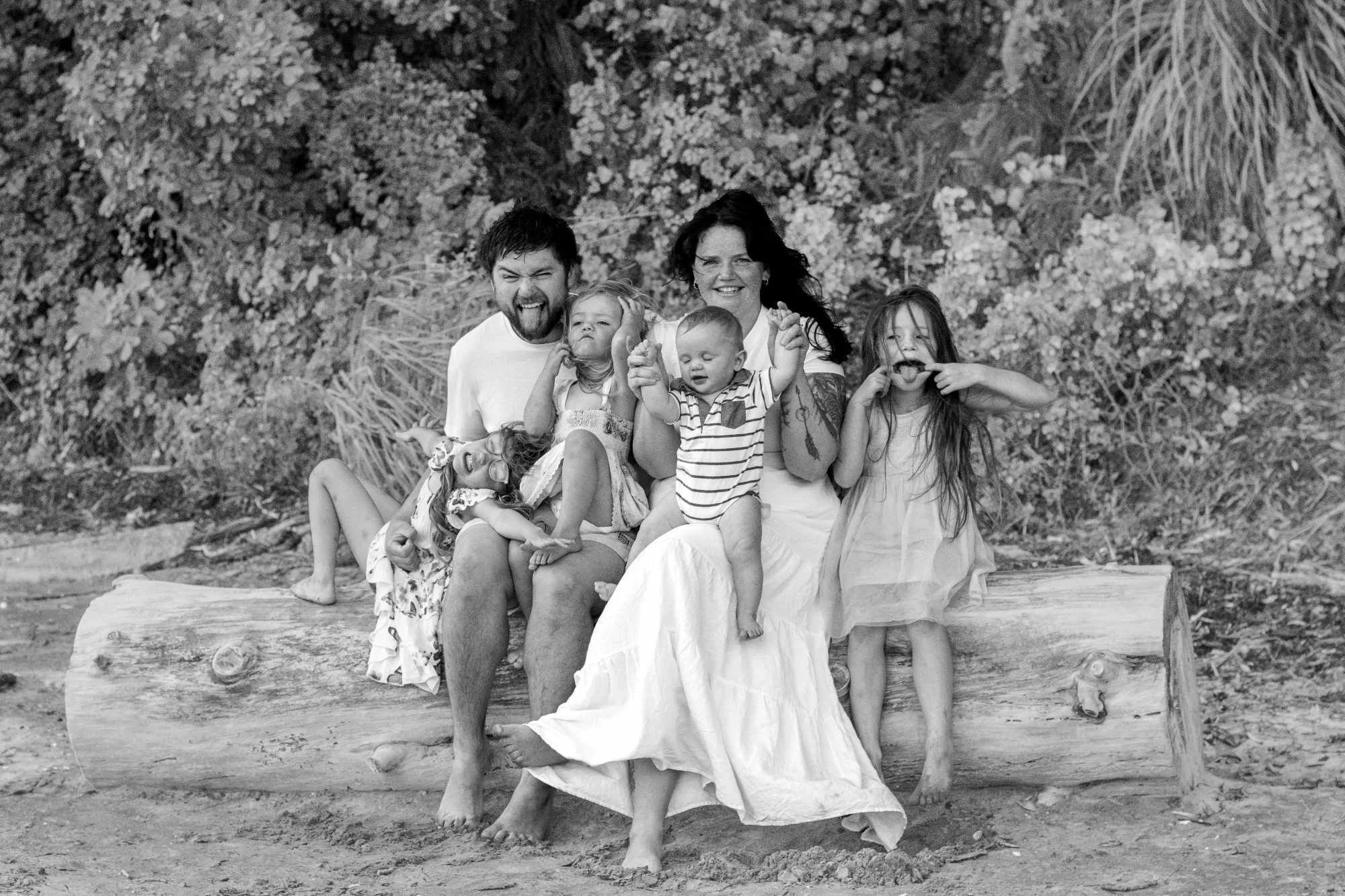 A family of six posing on a large fallen log outdoors, surrounded by bushes and trees, black and white photograph.