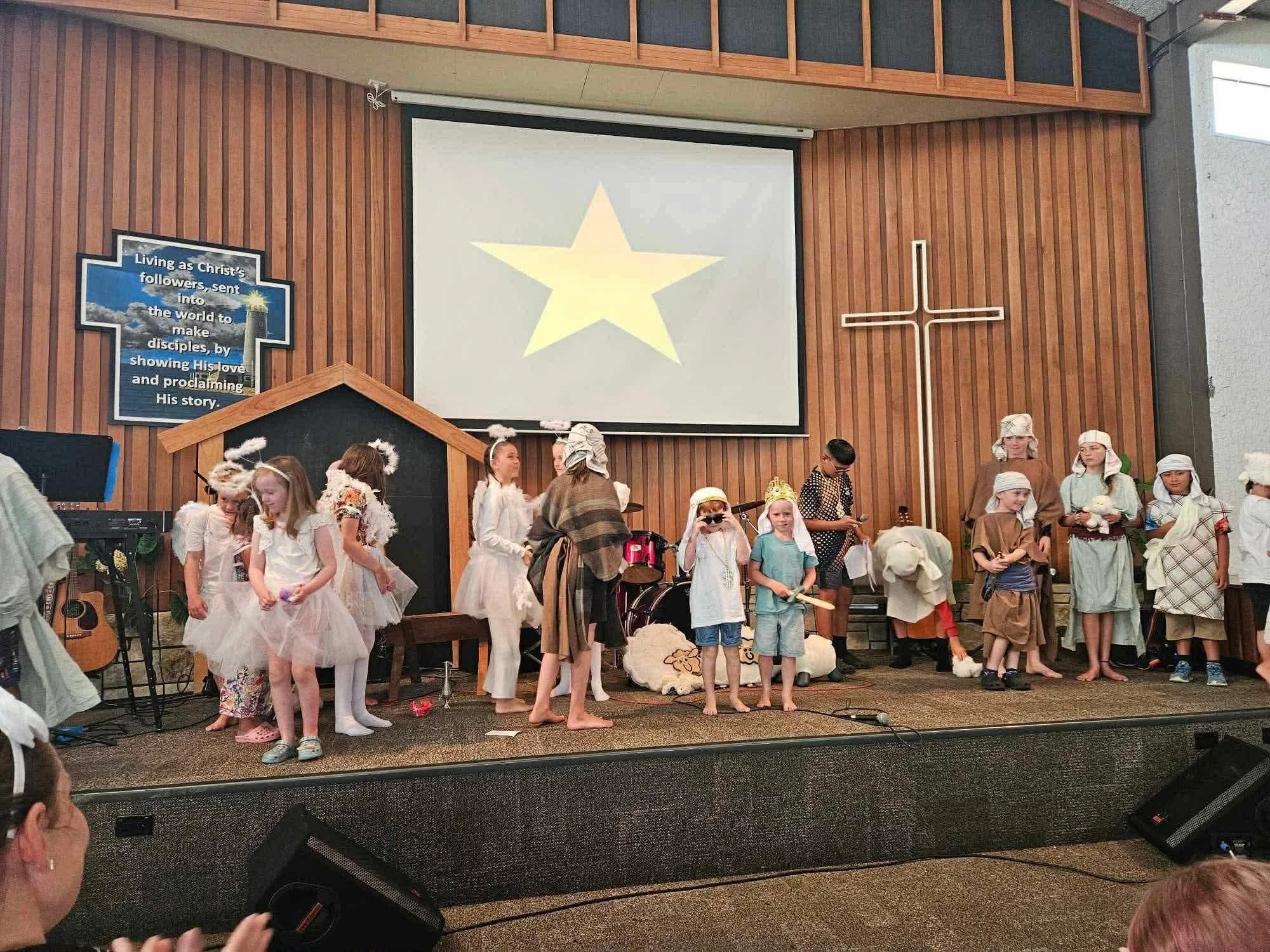 Children dressed as angels and shepherds performing a Christmas play on stage inside a church. A cross and a large star are displayed on the wall behind them.