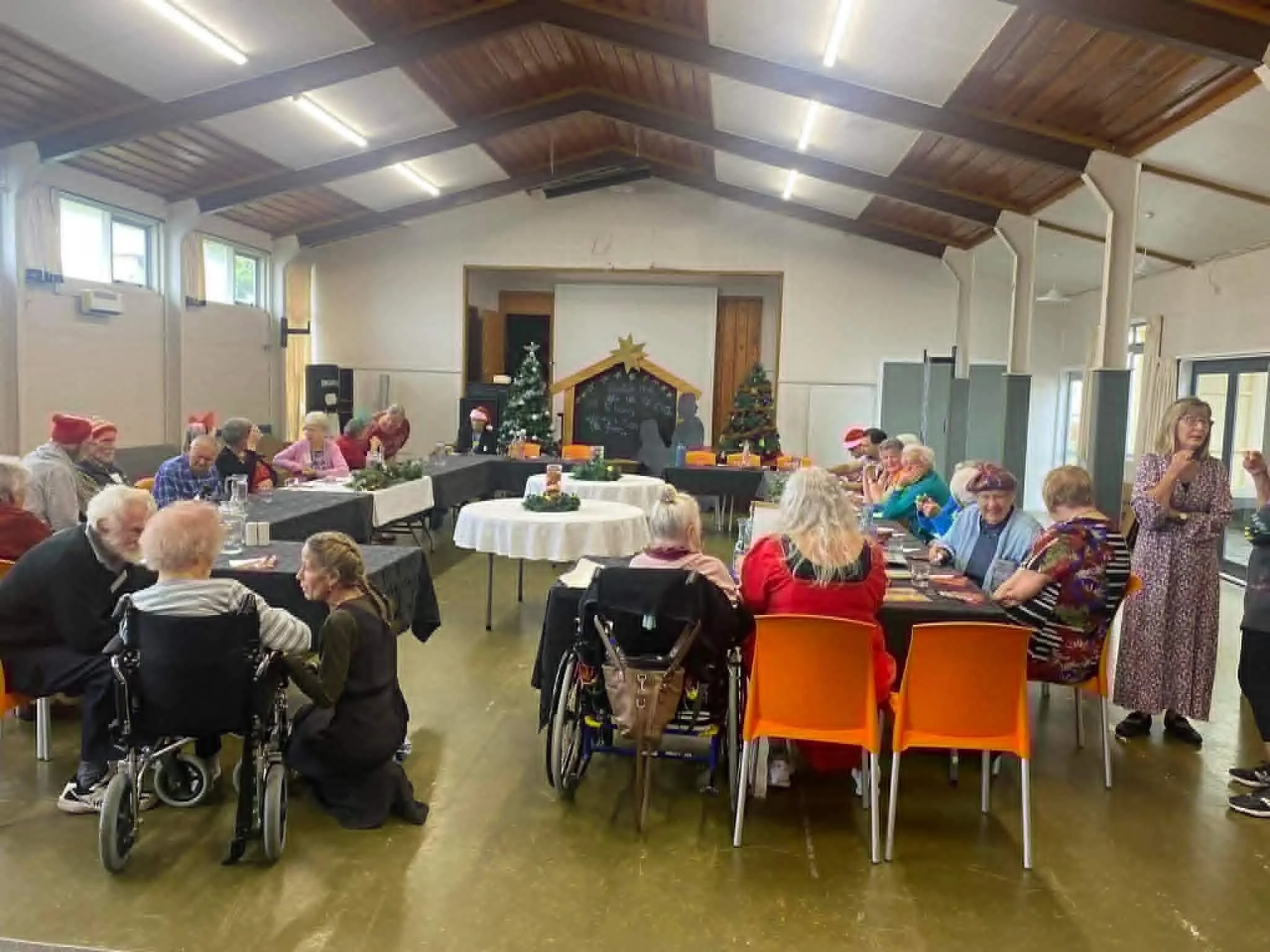 People gathered in a decorated church hall for a Christmas celebration, with Christmas trees, festive decorations, and people wearing Santa hats.