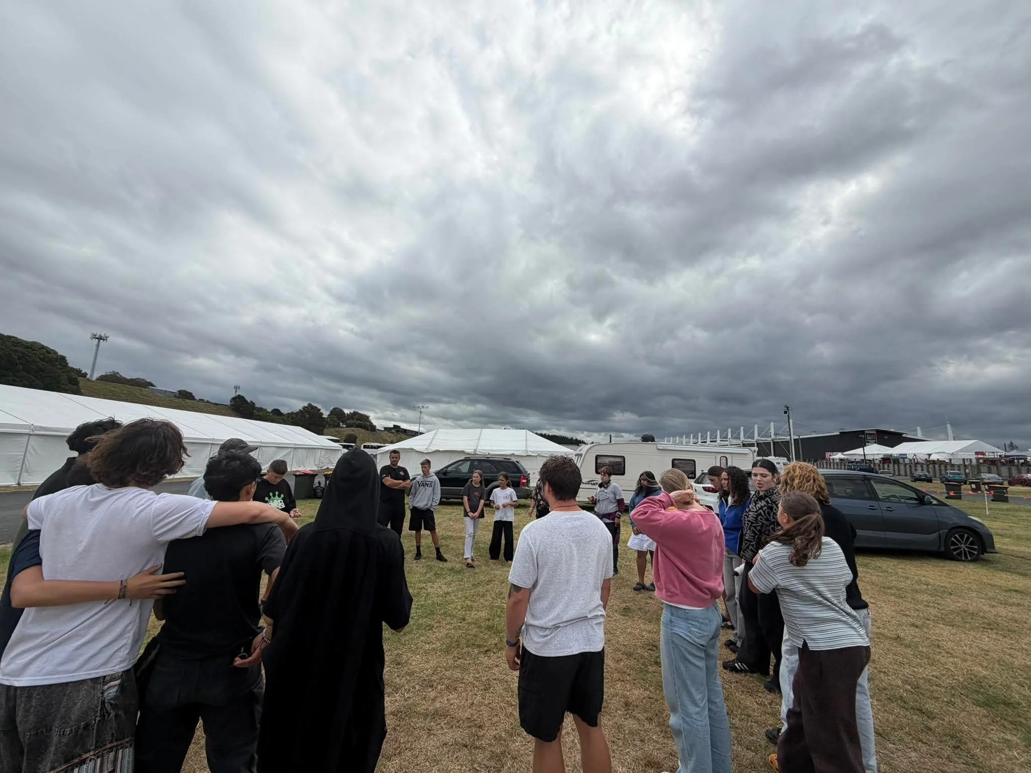 A group of people standing in a circle outdoors on grassy ground in front of white tents and vehicles, under a cloudy sky.