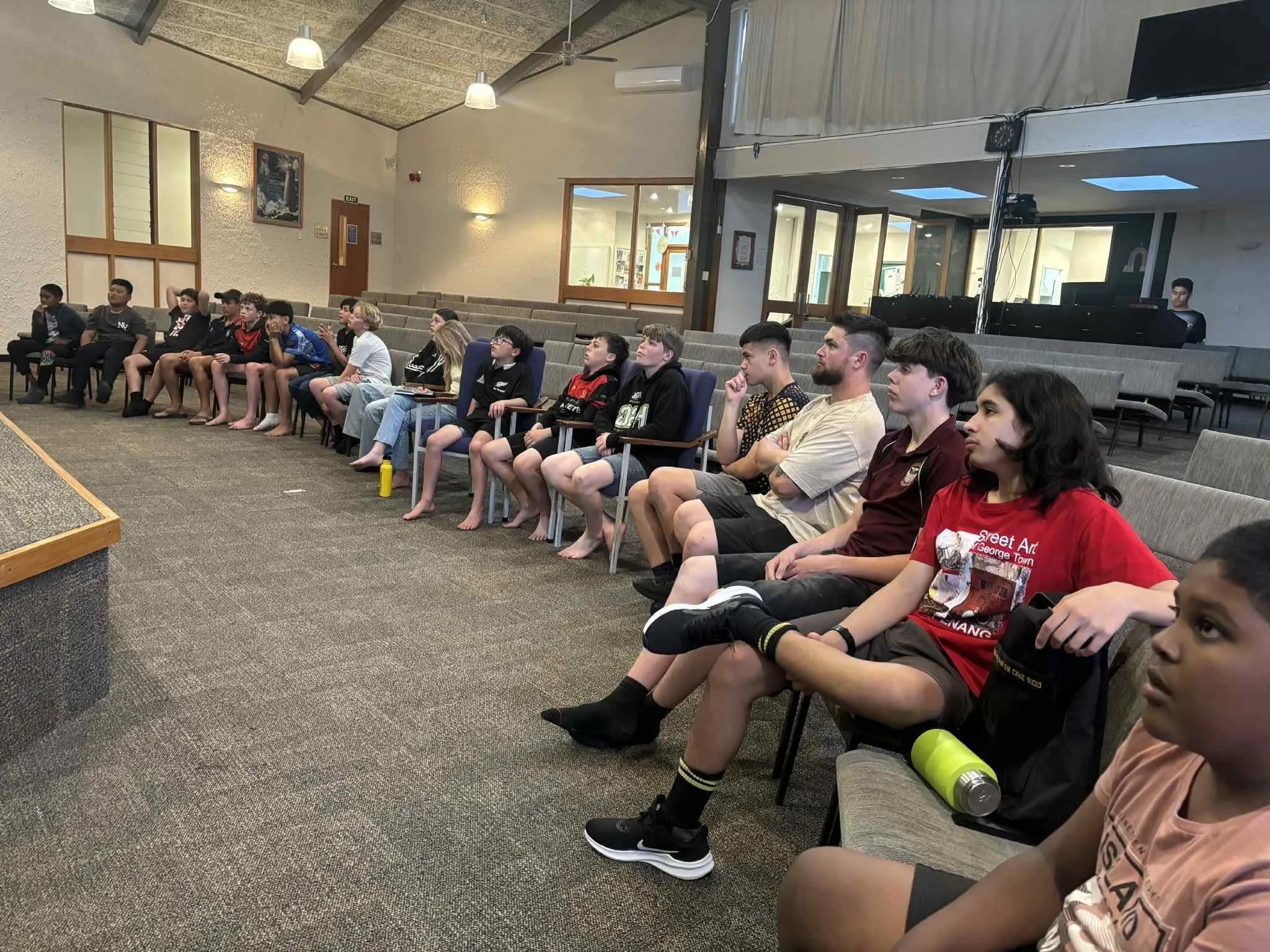 Group of young students and adults seated in a semi-circle in a room, watching an unseen presentation or performance.