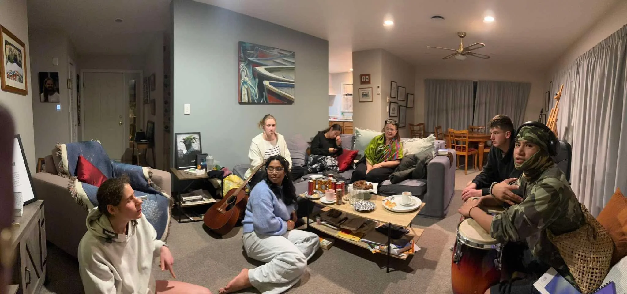 People gathered in a living room for a casual get-together. There are seven individuals, some sitting on couches and chairs, with snacks, drinks, and a guitar visible on the table and floor. The room features wall art, framed photos, and curtains covering large windows.