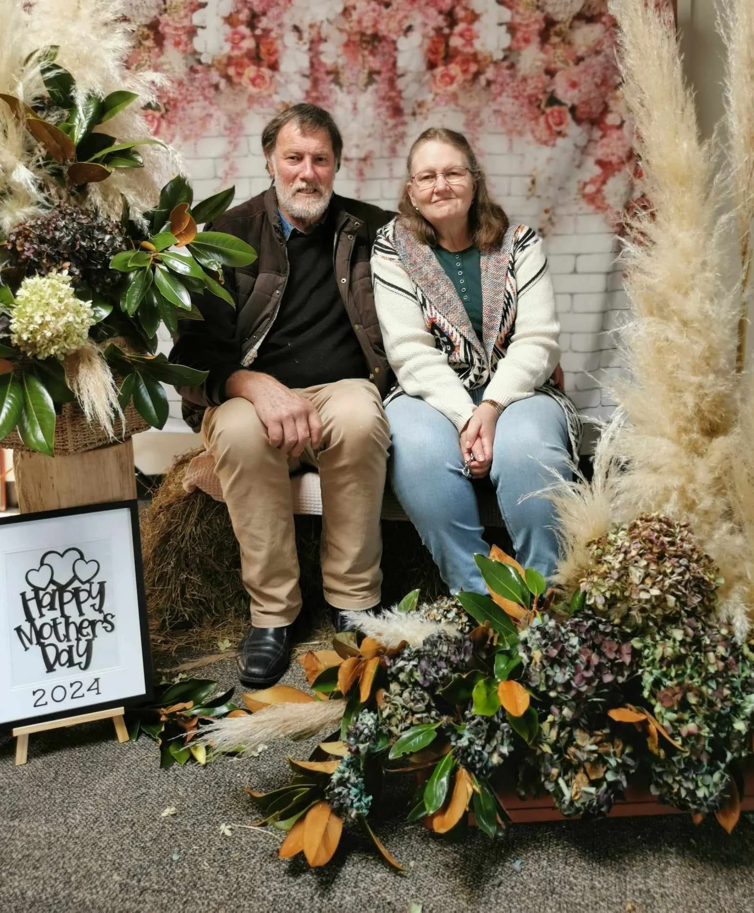 A man and woman sitting on a small bench surrounded by flower arrangements and pampas grass, with a 'Happy Mothers Day 2024' sign beside them.