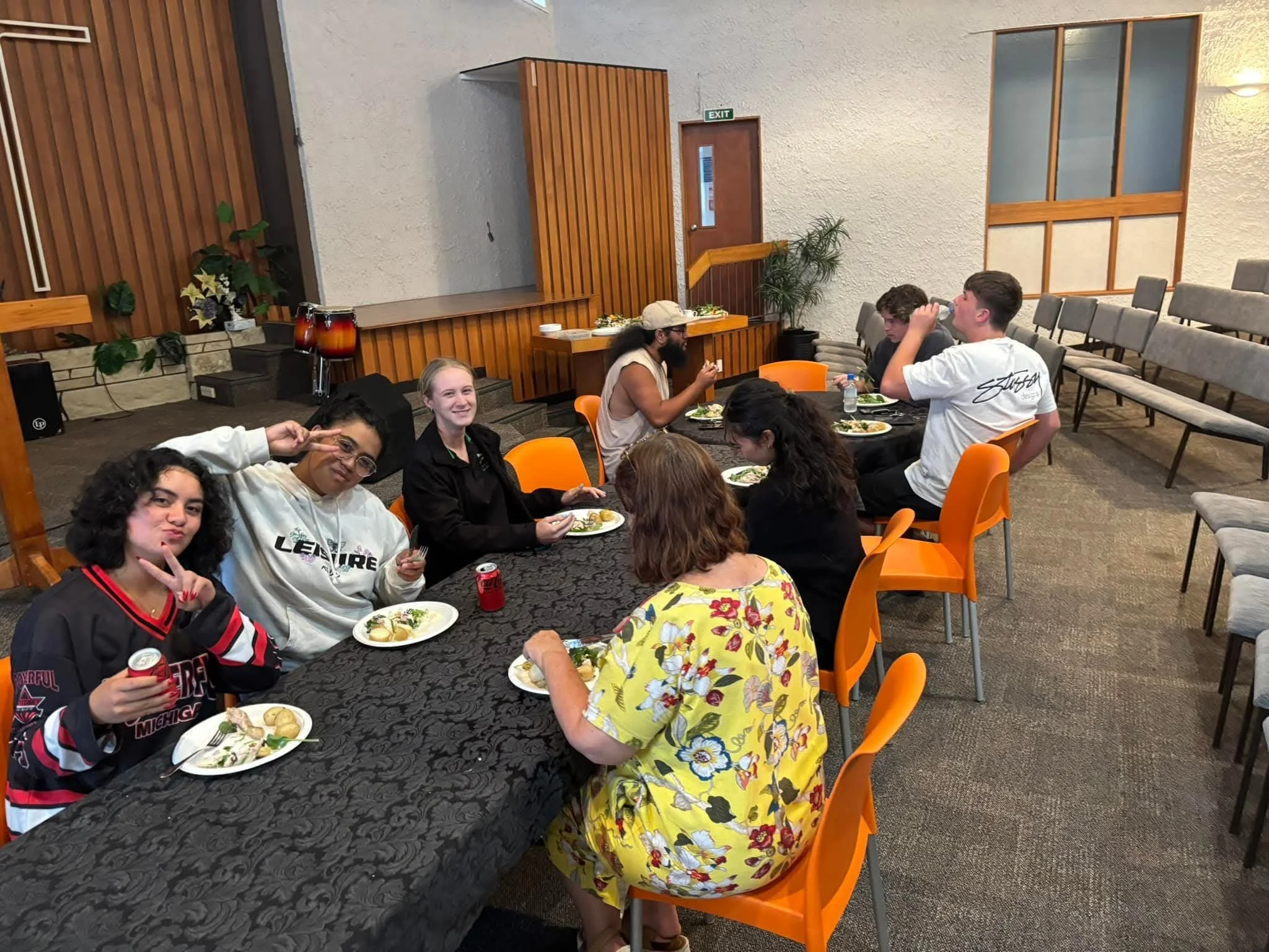 A group of nine people sitting at a long table in a banquet hall, enjoying a meal together. The room has a wooden wall, a potted plant, and a stack of chairs against the wall.