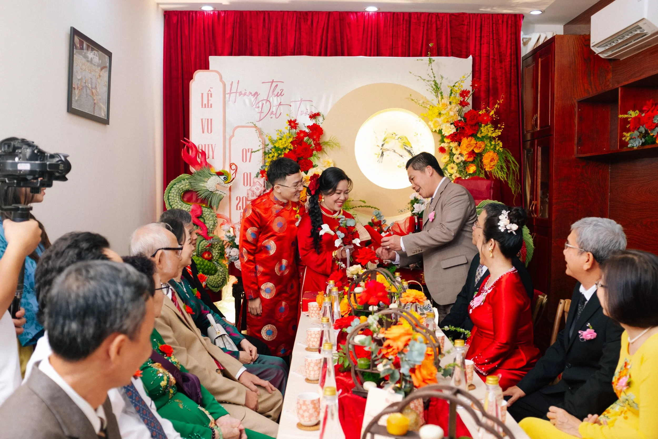 Vietnamese wedding banquet with a bride and groom exchanging vows, surrounded by family and friends dressed in traditional and formal attire, decorated with vibrant flowers and red accents.