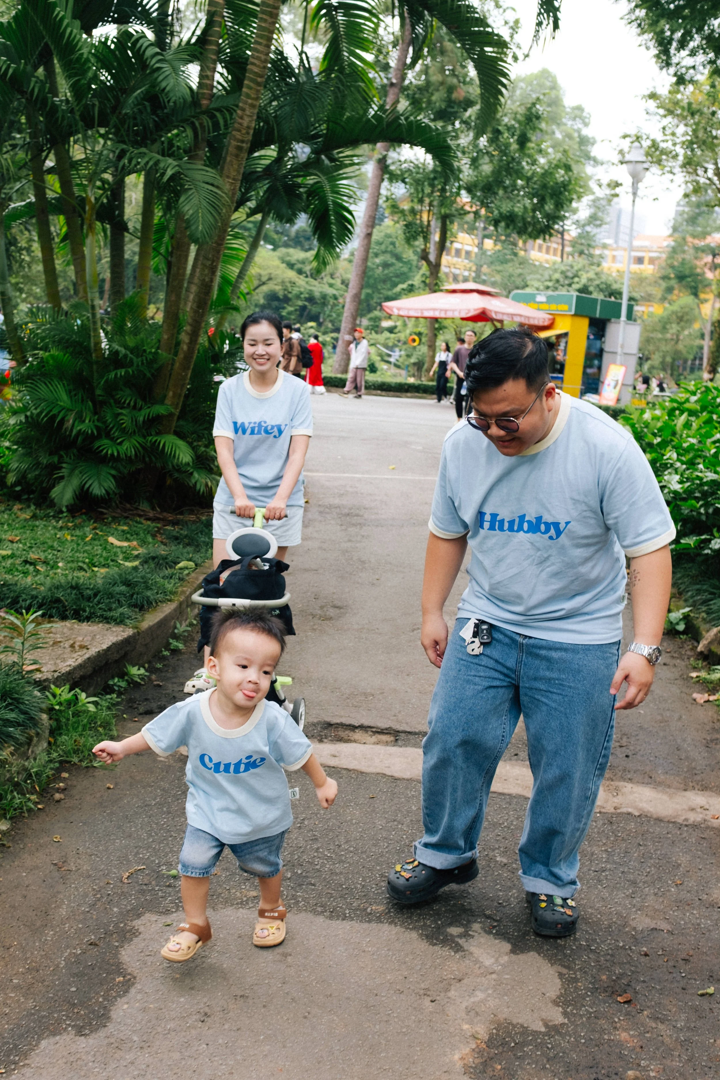 A group of people, including a young child, a woman, and a man, walking in a park. The child is running ahead, while the woman pushes a stroller, and the man is smiling and looking down. They are all wearing matching light blue shirts with words like