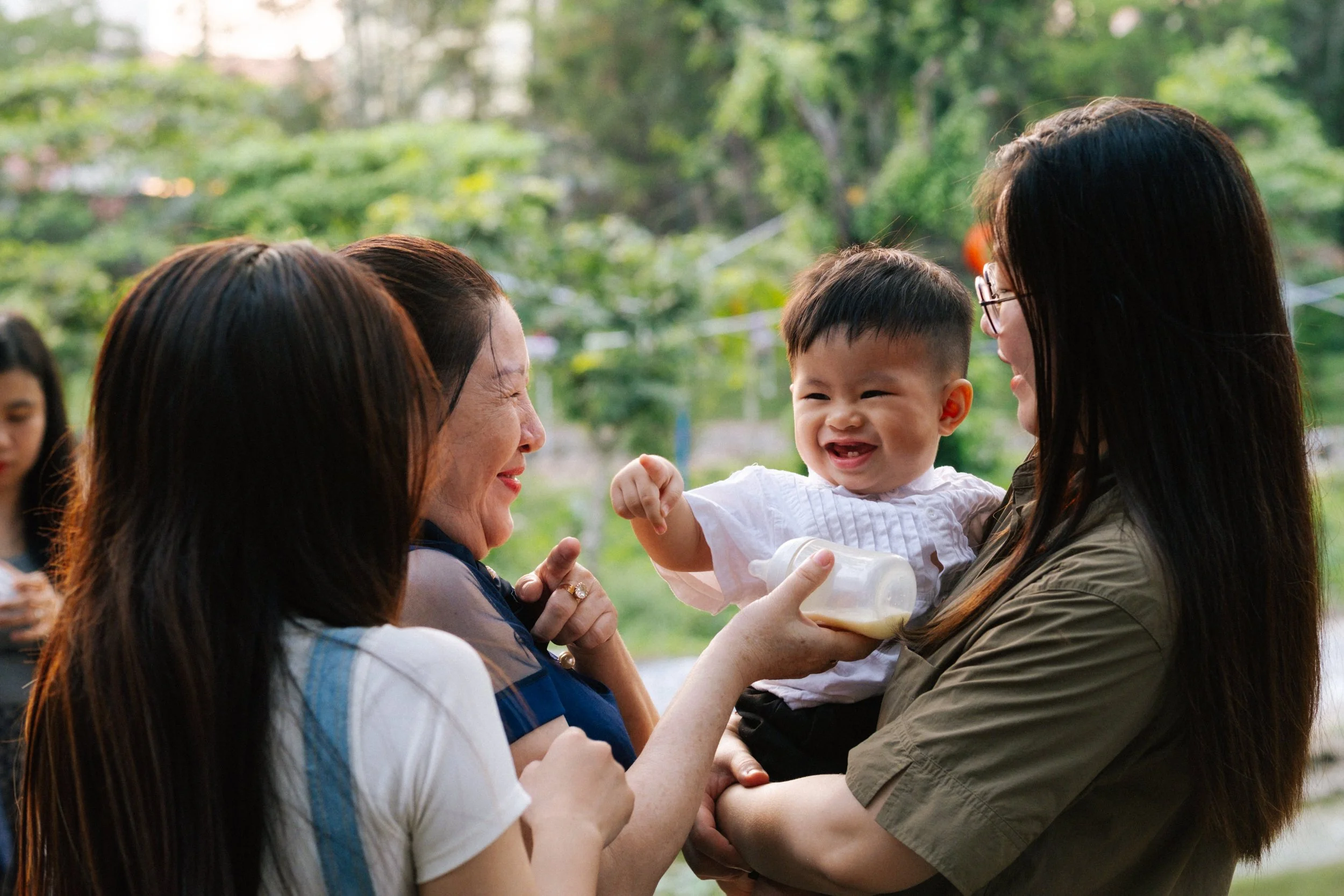 A group of women and a baby smiling and interacting outdoors in a green park during daytime.