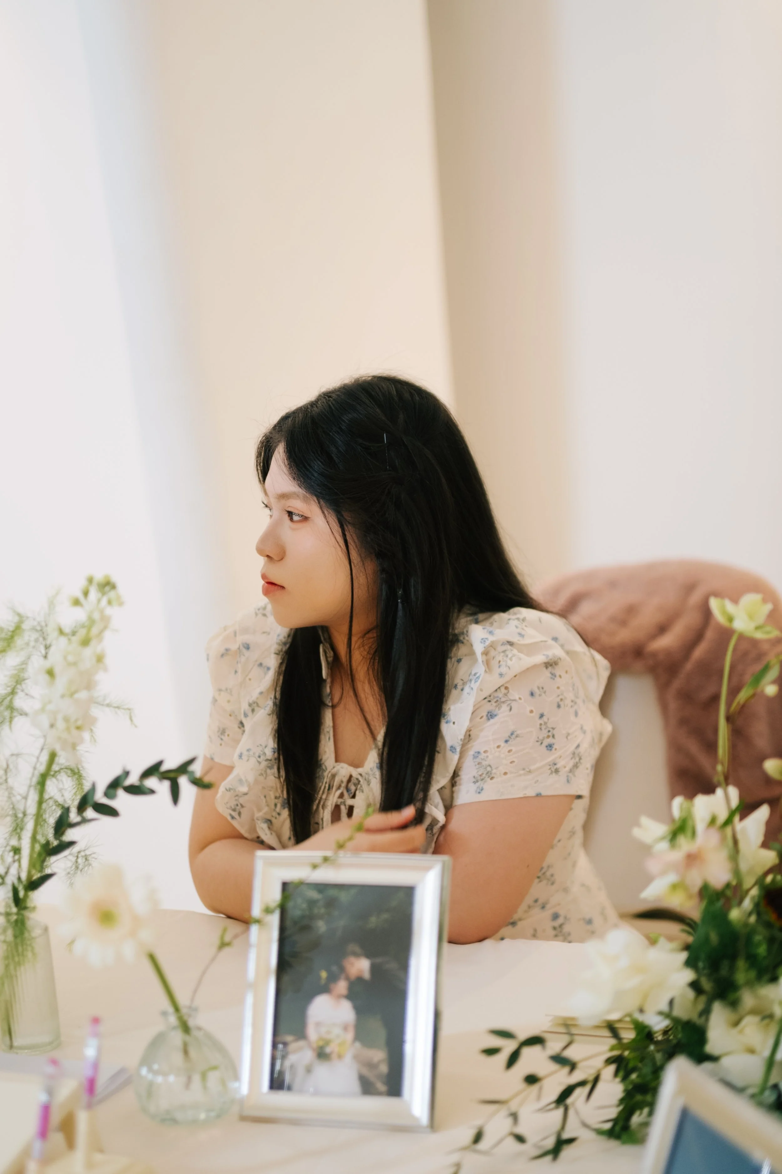 A woman with long black hair and light skin is sitting at a table decorated with flowers and framed photographs, looking to her left with a calm expression.
