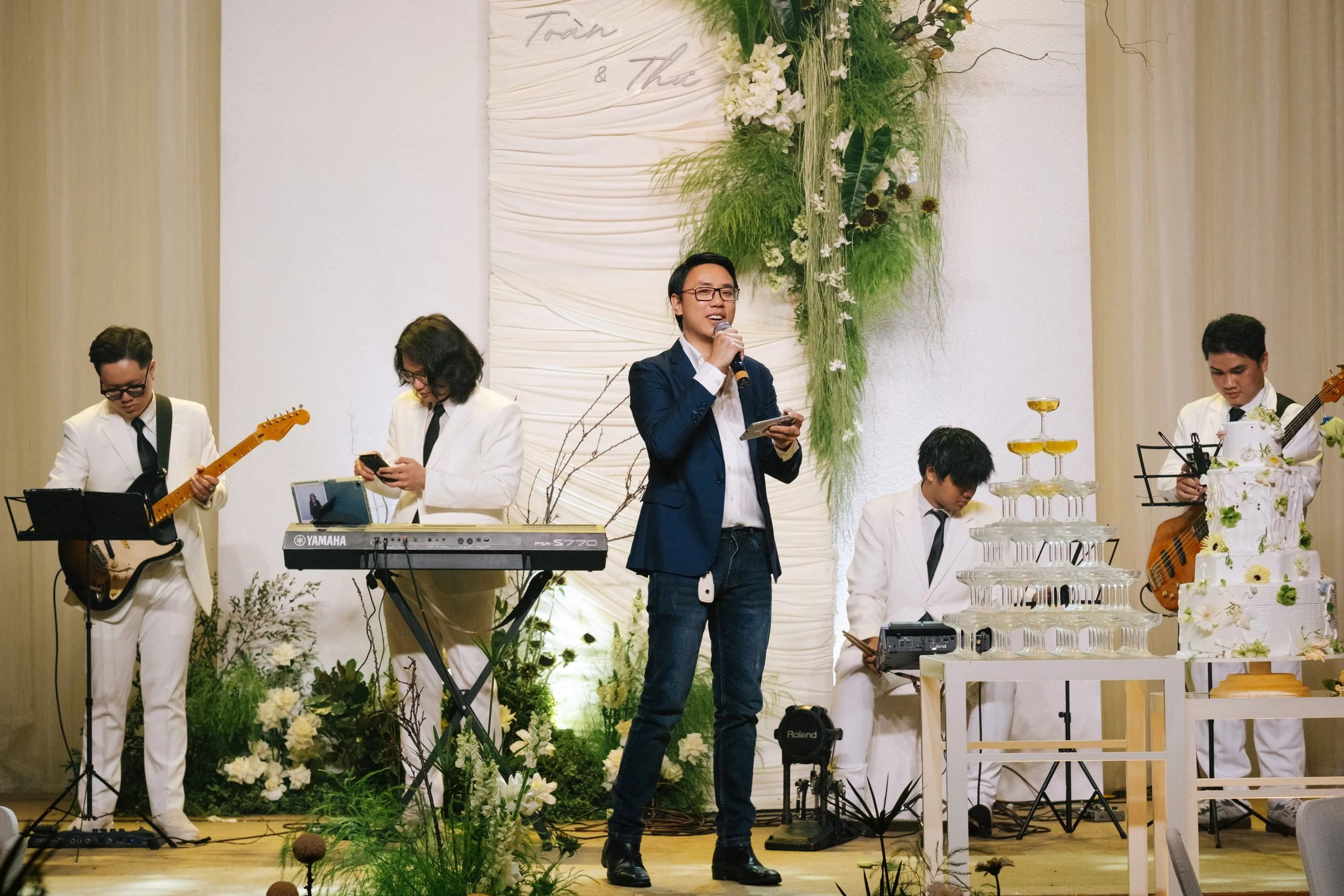 A man in a blue blazer and jeans speaking into a microphone at a wedding or event, surrounded by musicians dressed in white suits playing guitar, keyboard, and bass, with a multi-tiered wedding cake and decorative greenery in the background.