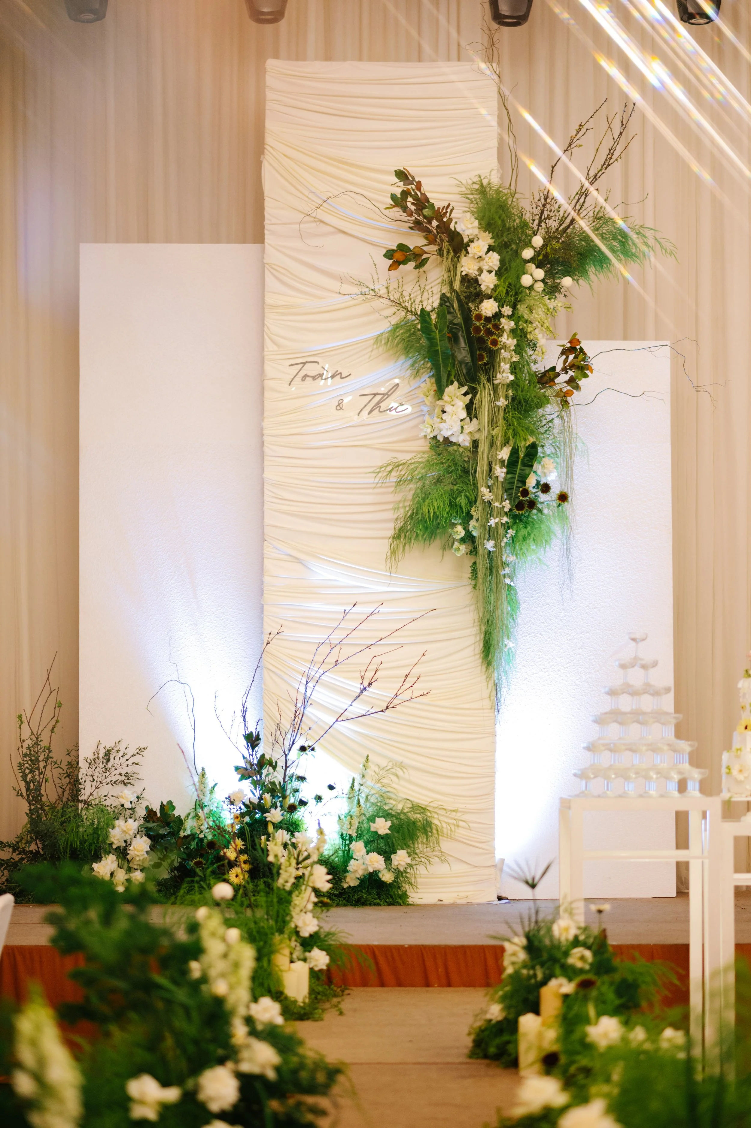 A wedding altar decorated with white and green flowers and foliage, featuring a large vertical fabric backdrop with a handwritten sign, illuminated with soft lighting.