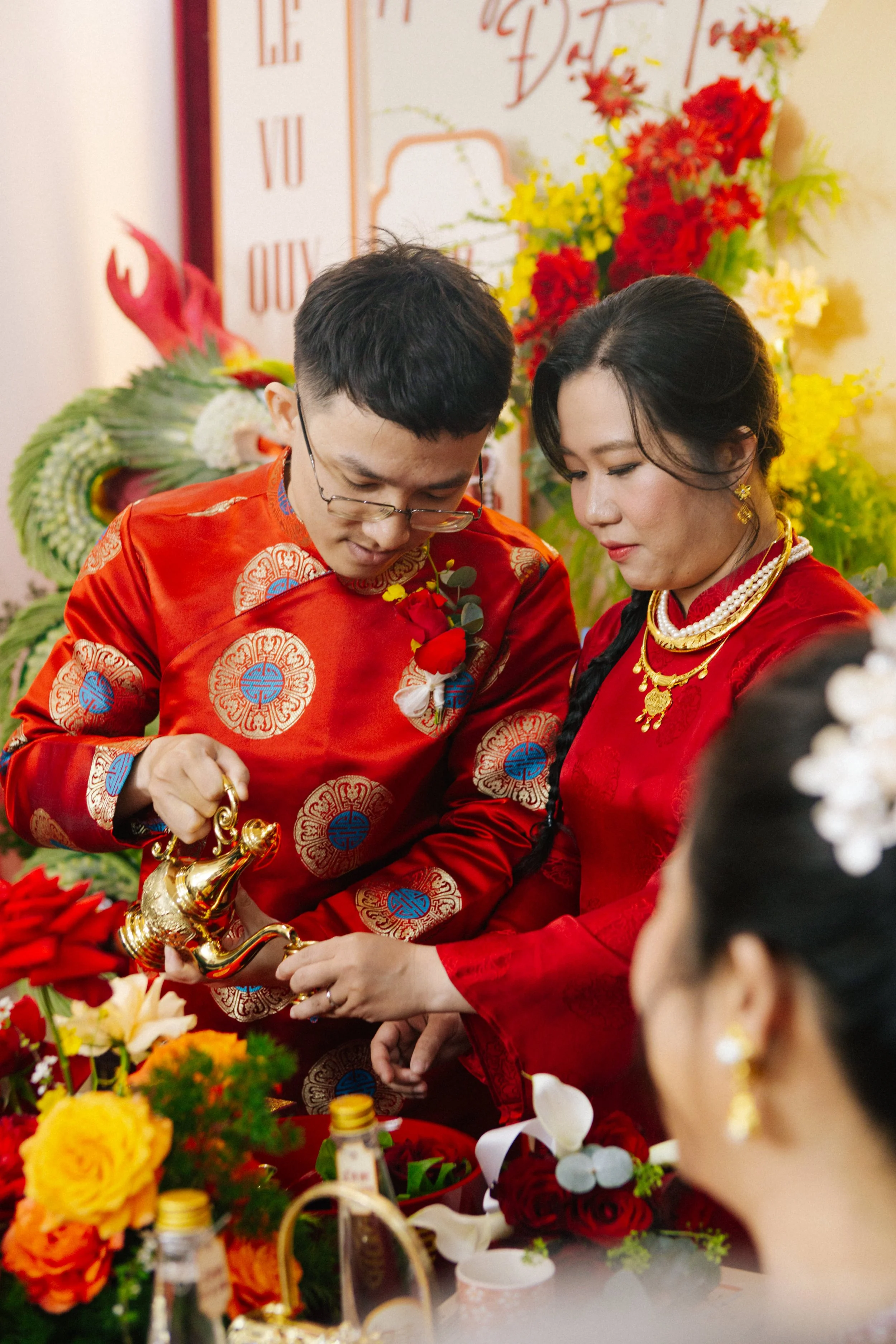 A couple in traditional red Vietnamese wedding attire participating in a tea ceremony, pouring tea from a gold teapot for a guest. The scene is decorated with colorful flowers and a backdrop with Vietnamese writing.