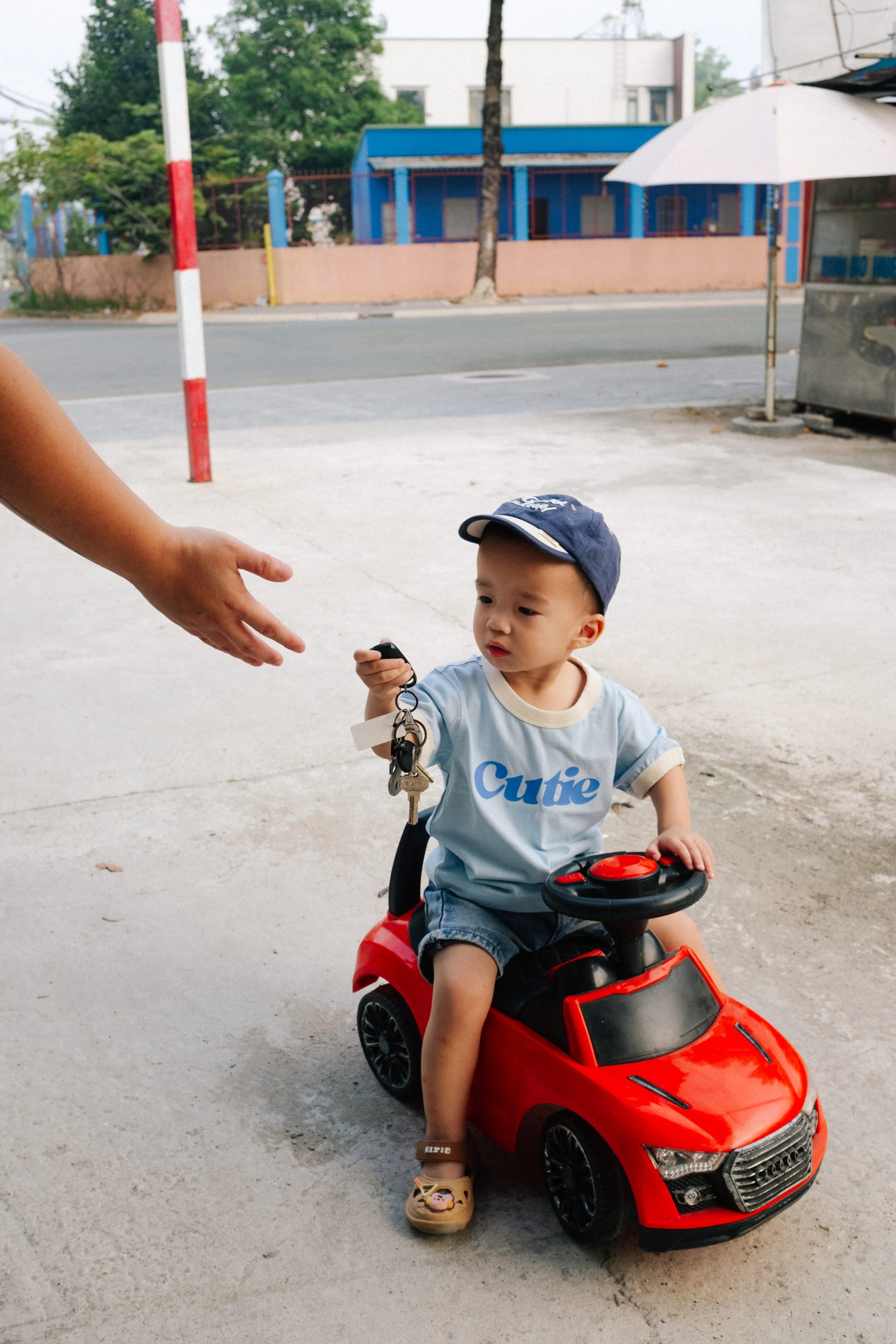A young boy riding a red toy car outside, holding a set of keys handed to him by an adult. The boy is wearing a blue cap, a light blue shirt with the word 'Cutie,' denim shorts, and beige shoes. There is an umbrella and some buildings in the backgrou