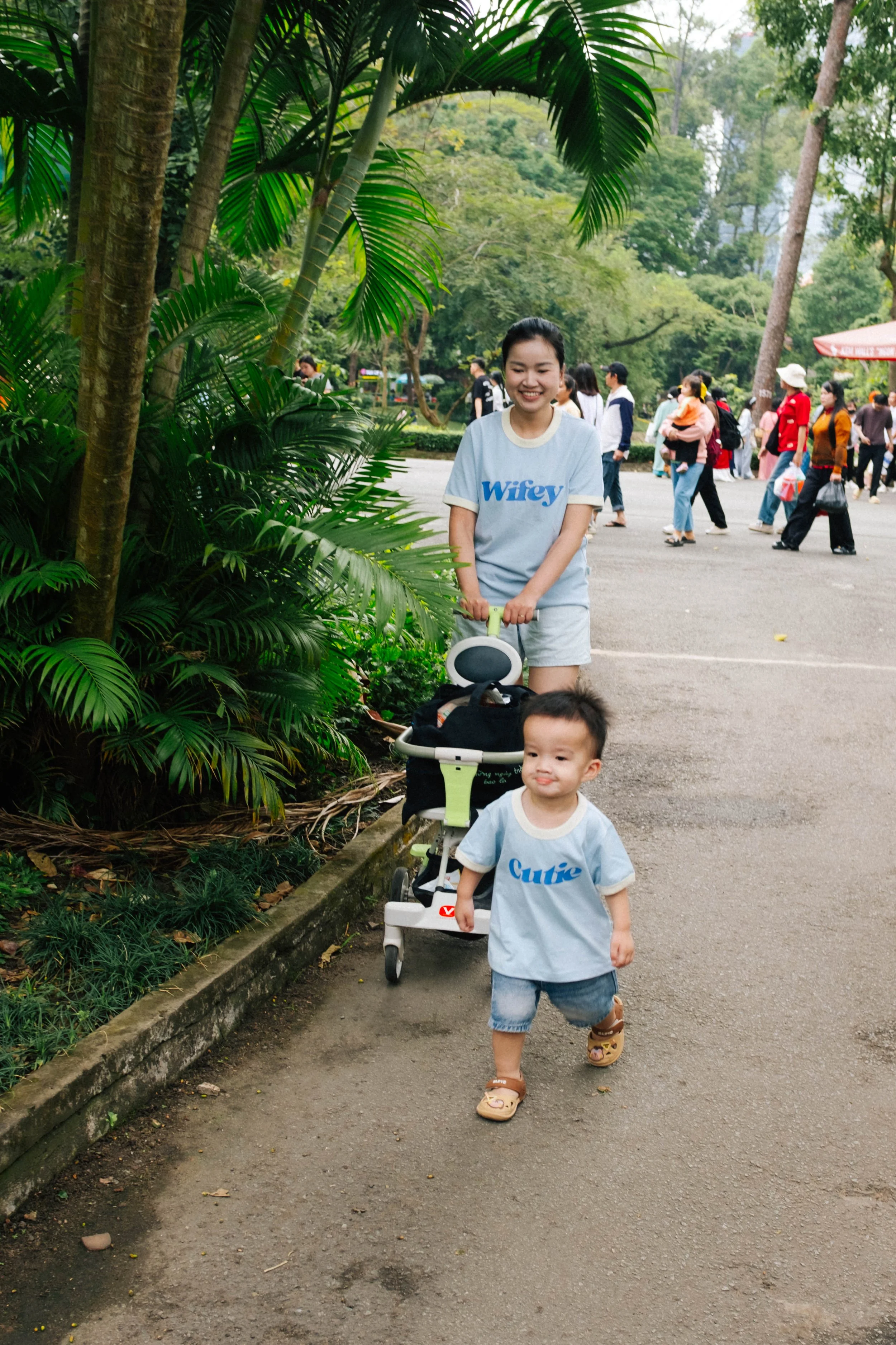 A woman and a young boy walking in a park, with the woman pushing a stroller. The boy is walking in front, wearing a blue shirt and shorts, while the woman is smiling and wearing a gray shirt with the word 'Wify' on it. There are many other people in