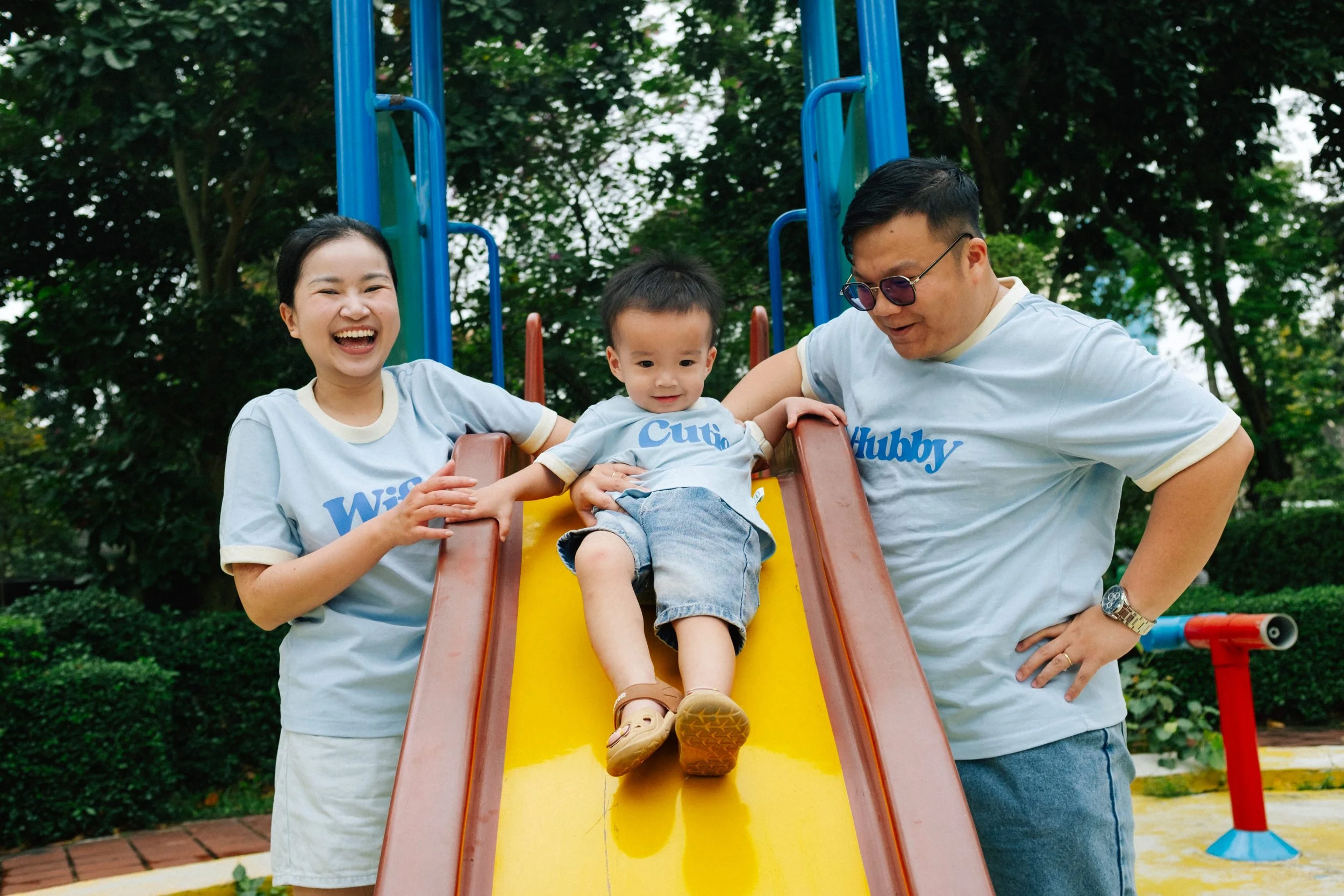 A happy family of three, a woman, a man, and a young boy, enjoying a slide at a park. They are wearing matching light blue t-shirts with funny words printed on them, and are surrounded by greenery.