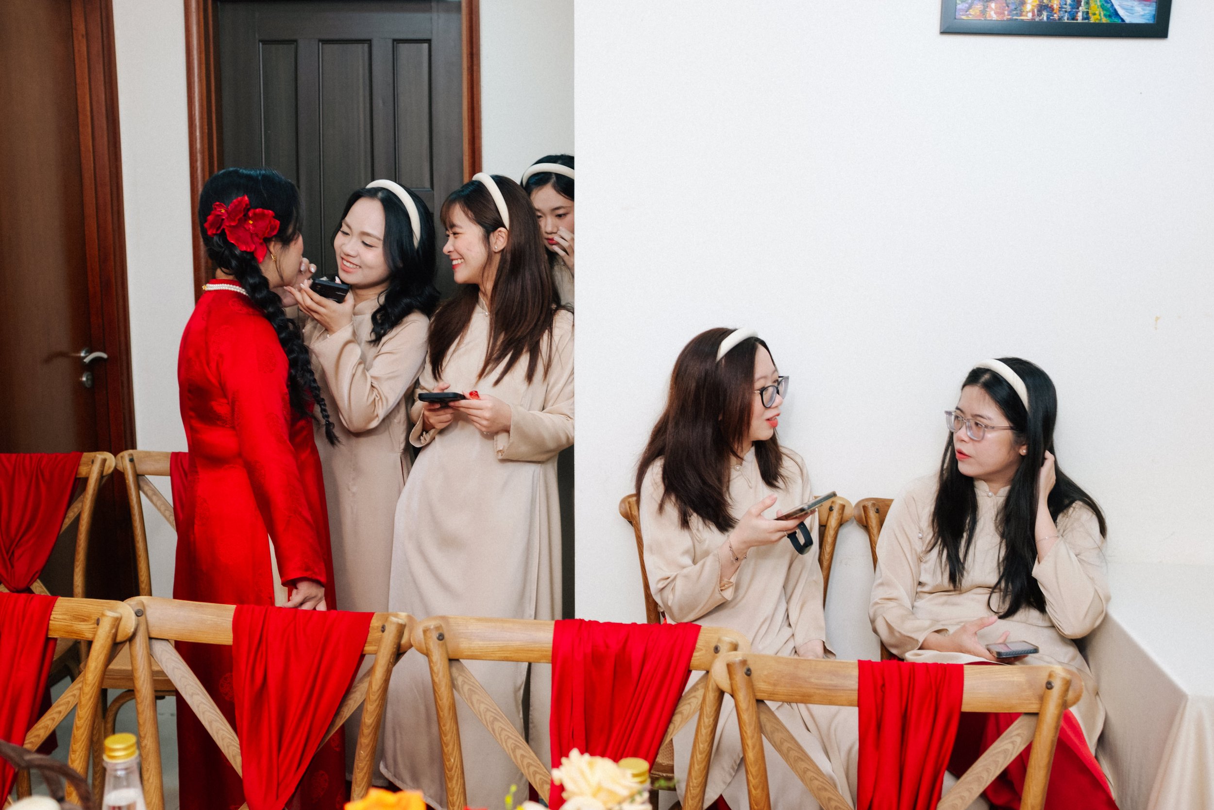 Women dressed in beige traditional dresses and headbands are gathered at a social event, with some sitting on wooden chairs with red fabric draped over the backrests, and one woman in a red traditional outfit with flowers in her hair standing in fron