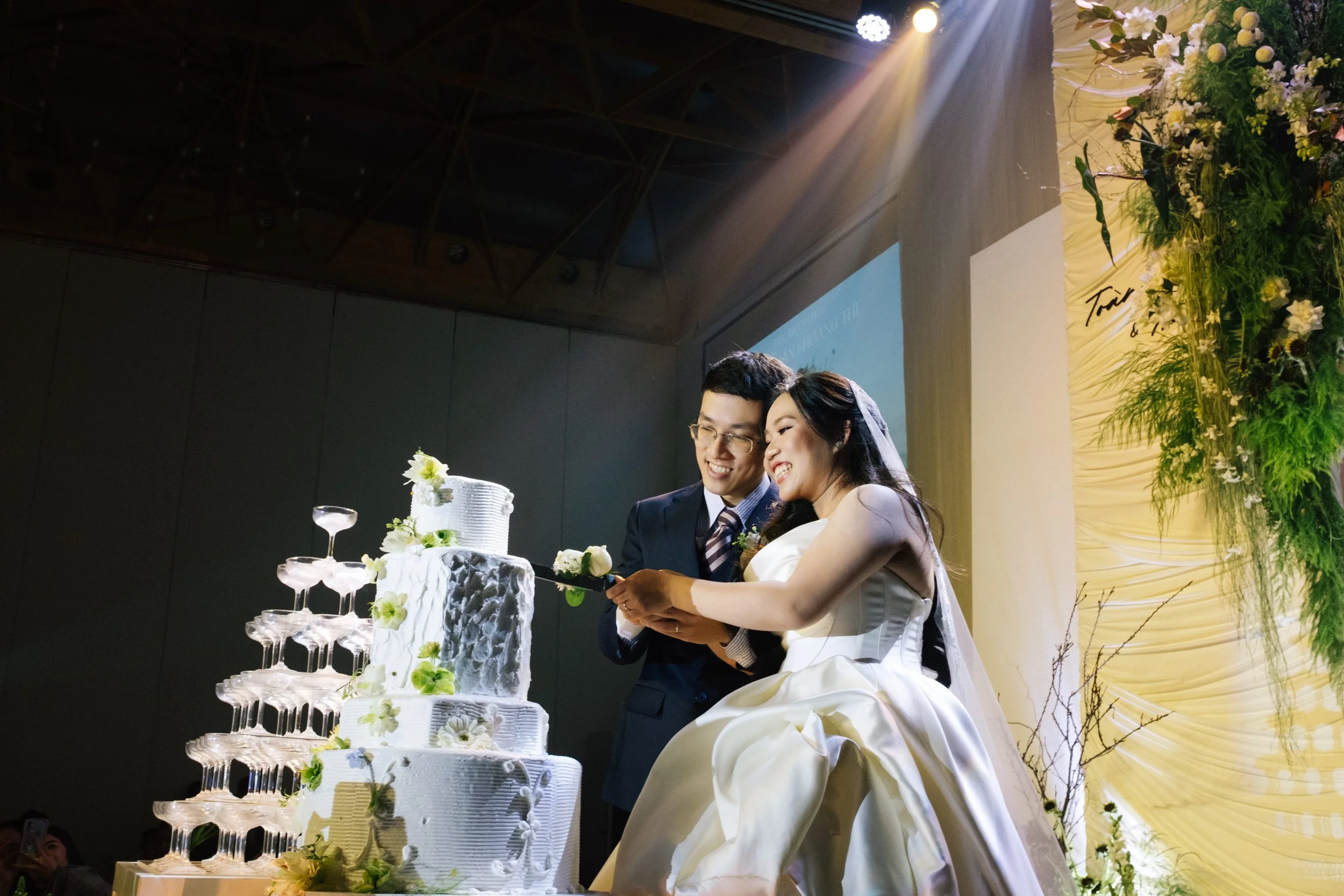 A bride and groom cutting a wedding cake together at their wedding reception, with the bride sitting in a wedding dress and the groom in a suit, smiling and enjoying the moment.