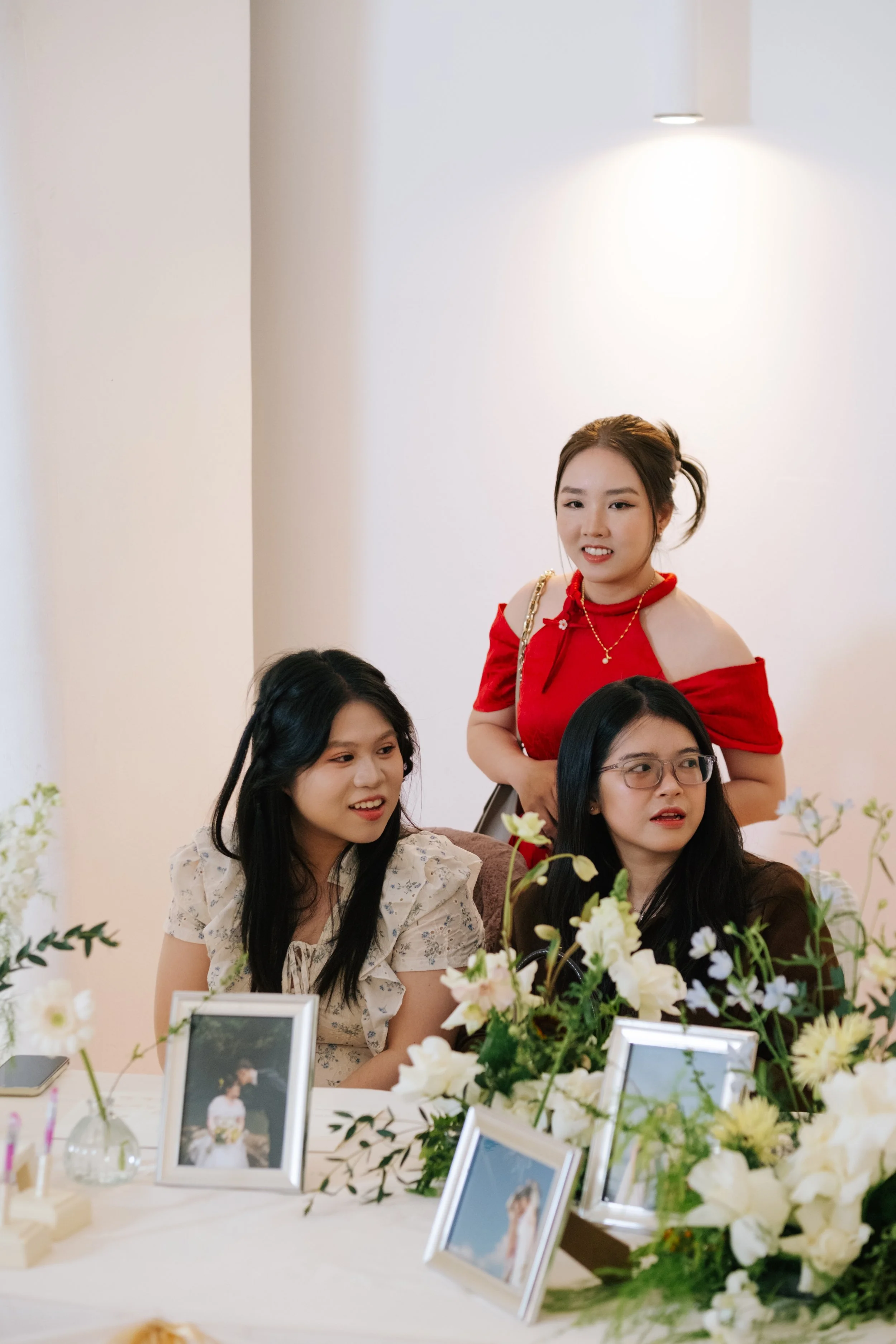 Three women sitting at a table decorated with flowers, with framed photos and a smartphone, during a social gathering or celebration.