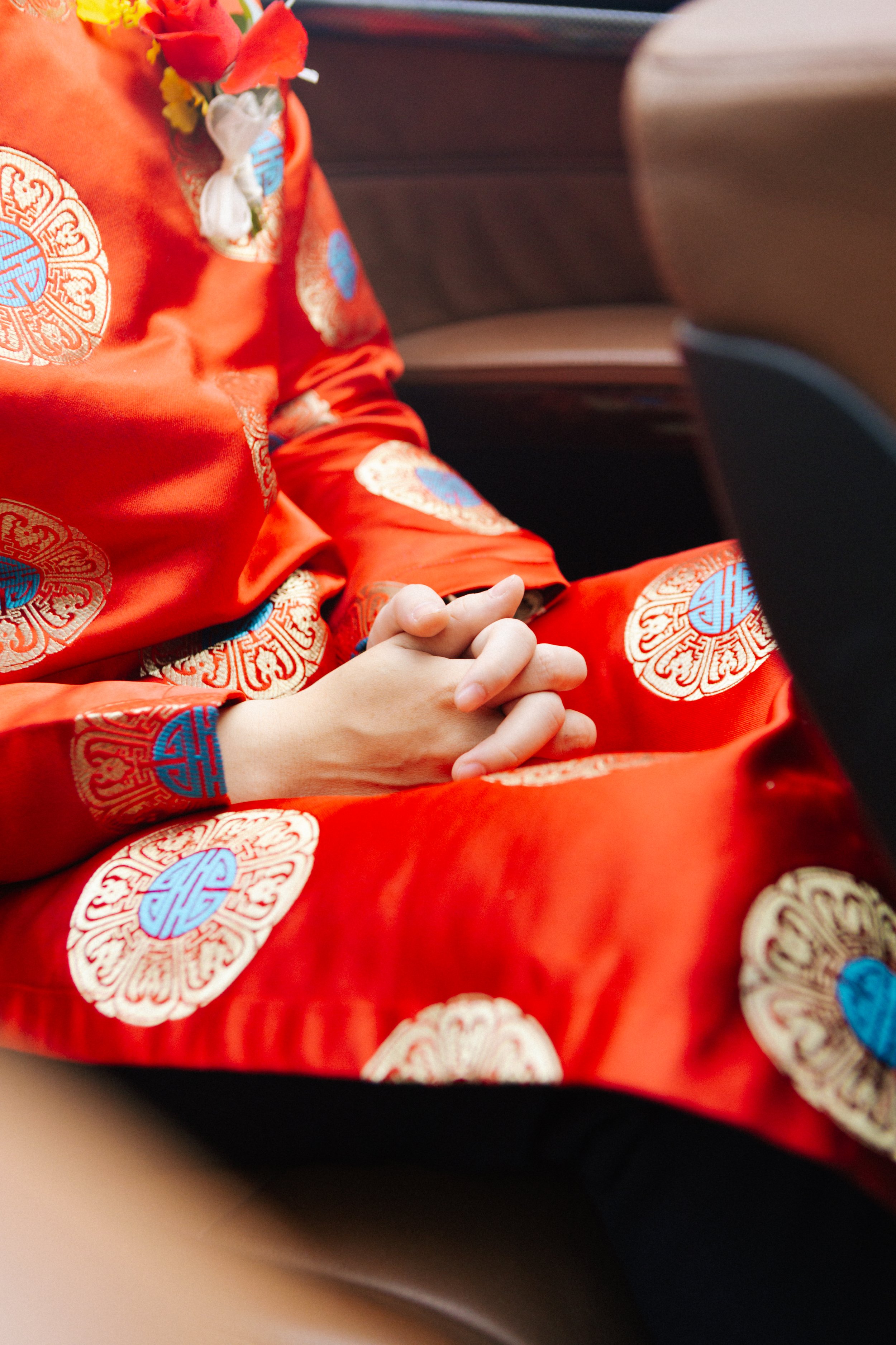 Person wearing a traditional red Chinese dress with gold and blue patterns, sitting with hands clasped together, sitting in a vehicle with brown leather seats.