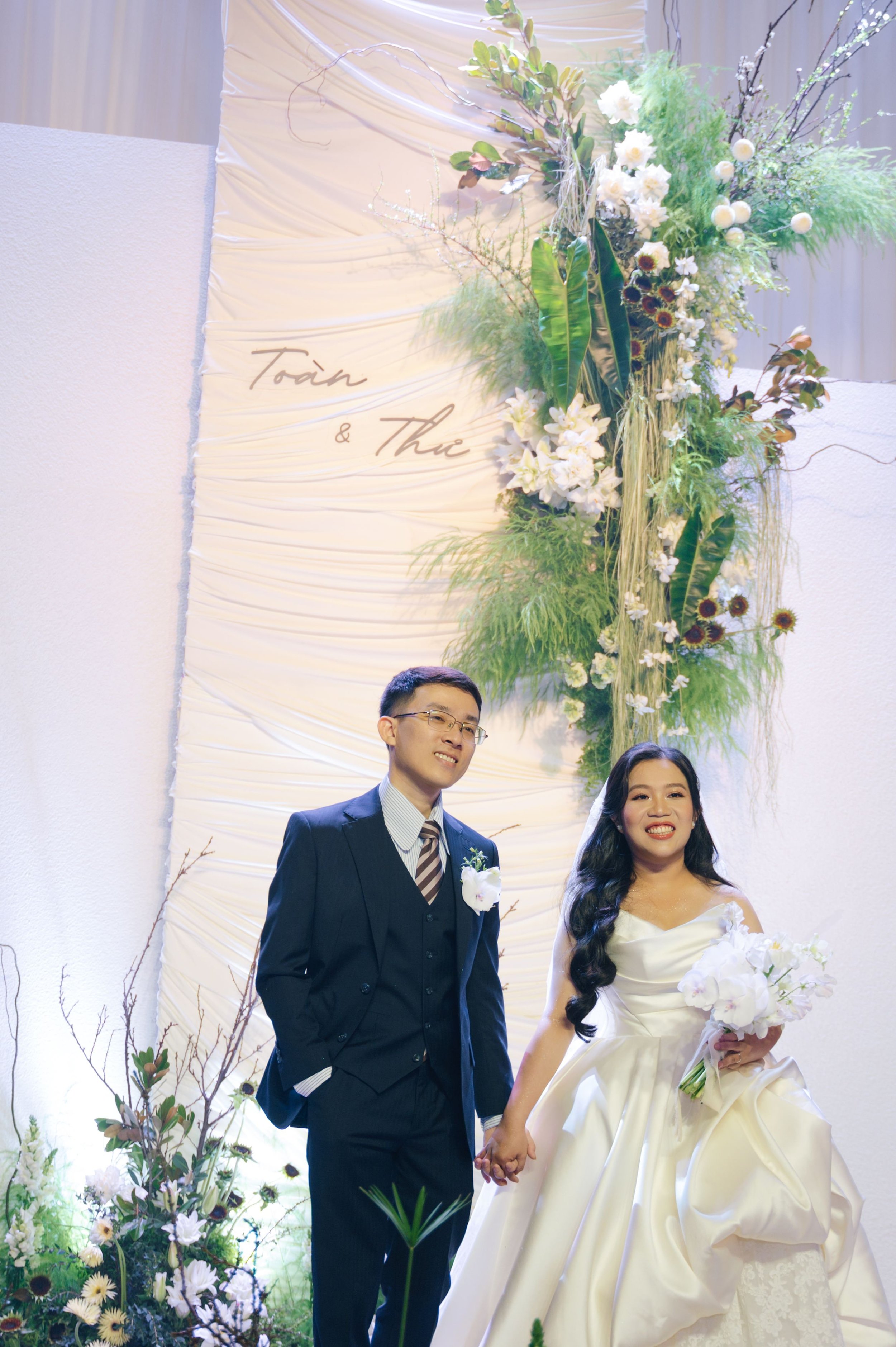 A bride and groom holding hands at their wedding, standing in front of a decorated backdrop with flowers and the words 'Toan & Thao' written on it.