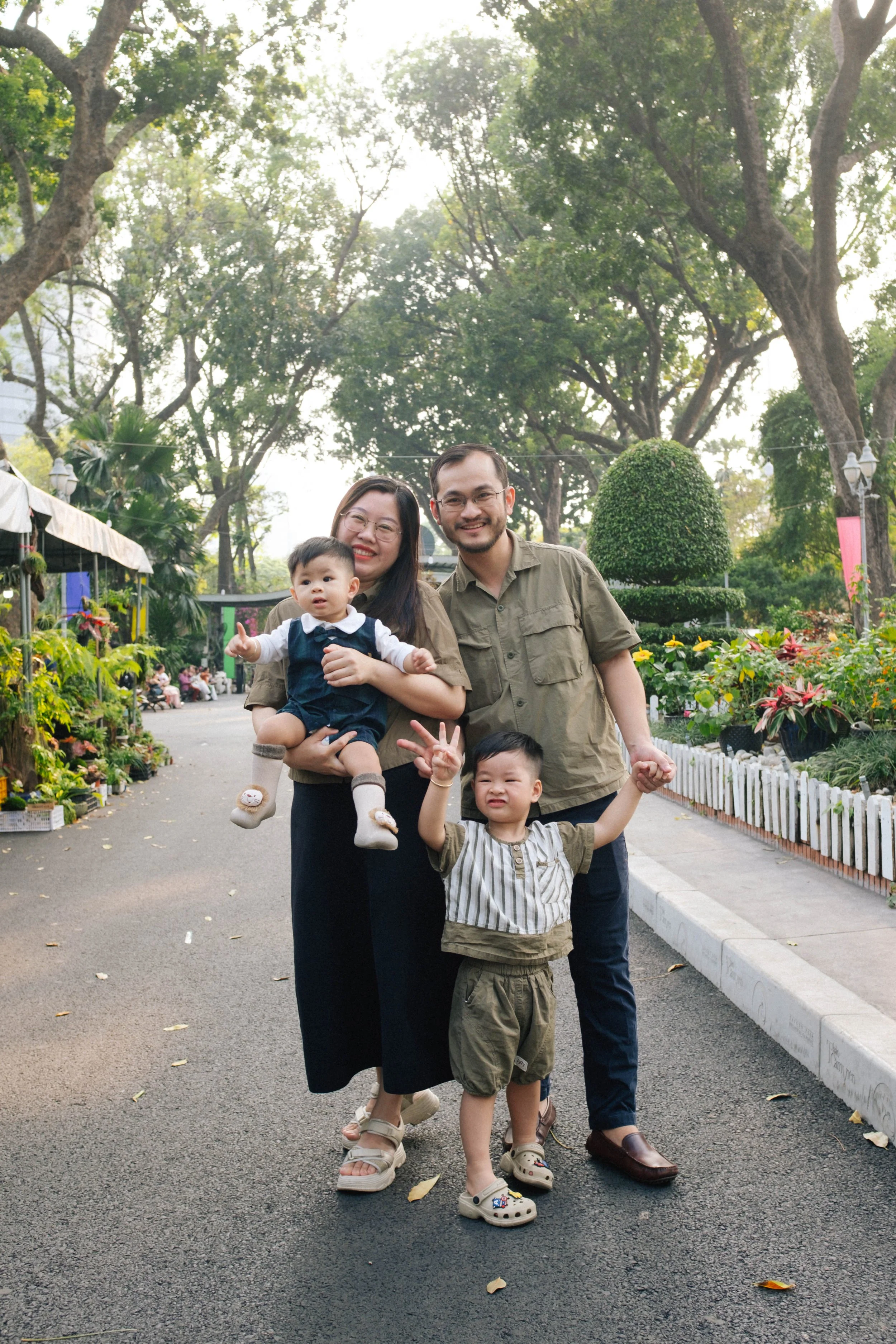 Family of four smiling and posing for a photo on a park pathway surrounded by greenery and flowers.