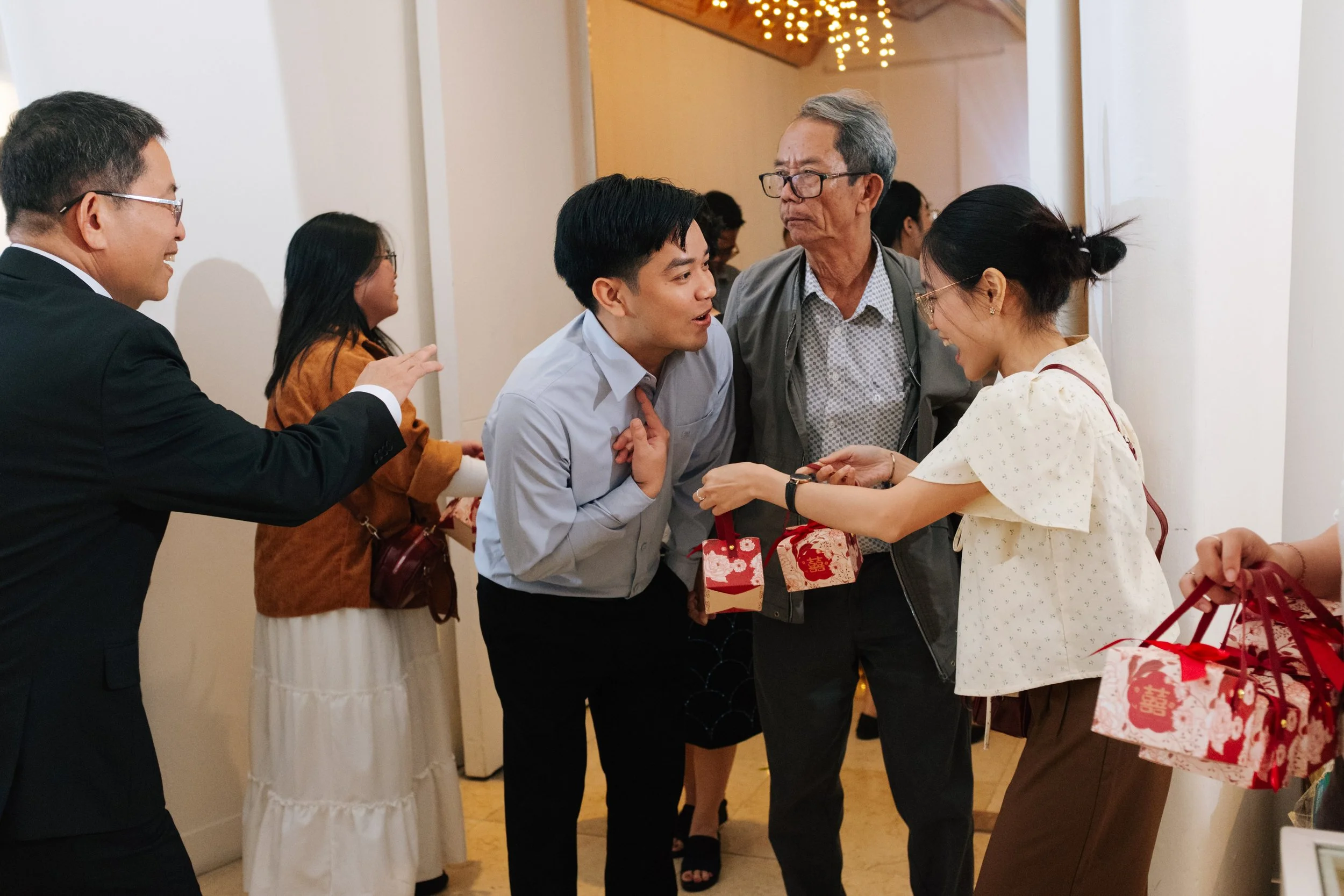 People interacting at a celebration, exchanging gifts, and smiling. A woman hands a gift to a young man, and others are nearby with more gifts and engaged in conversation.