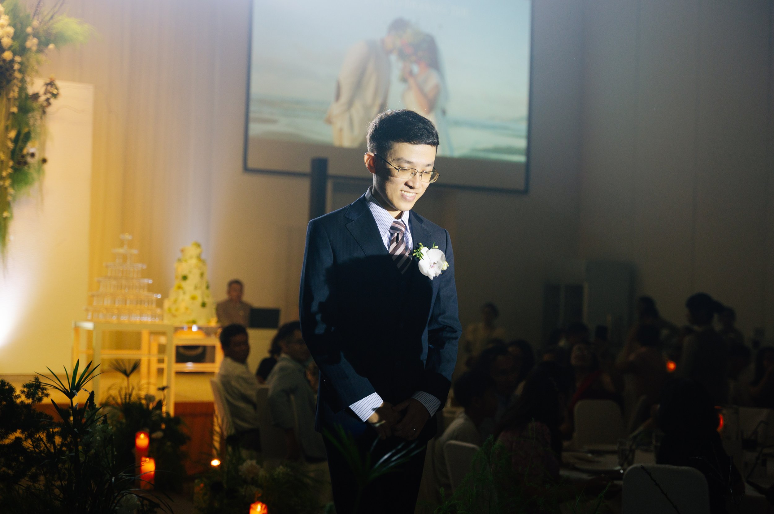 A young man in a suit with glasses and a white flower boutonniere stands in a dimly lit room at a wedding ceremony, smiling slightly and looking down. In the background, there are seated guests, floral decorations, and a large screen displaying an im