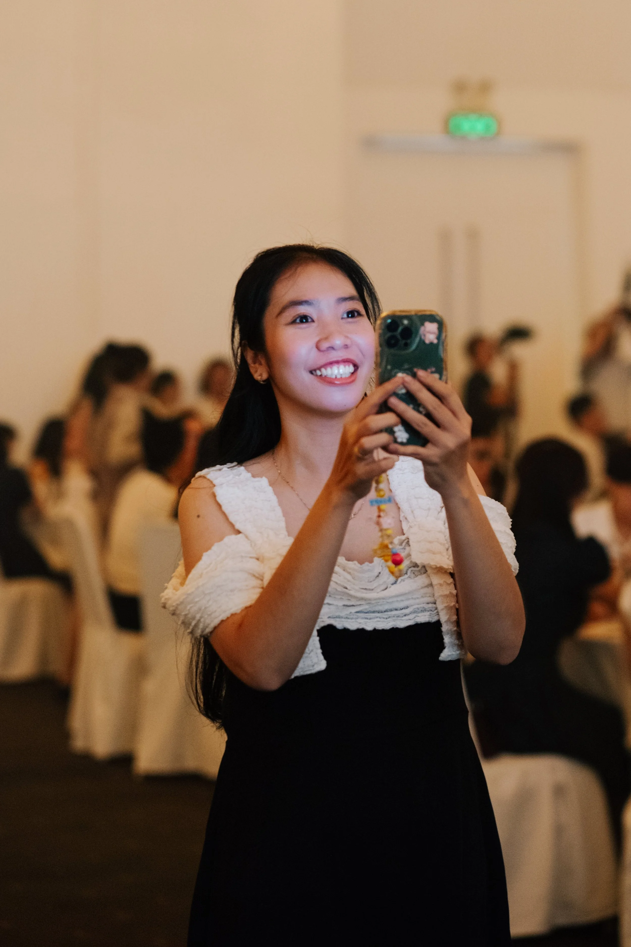 A smiling woman with long black hair taking a photo with her smartphone at a formal event or gathering, with other guests seated in the background.