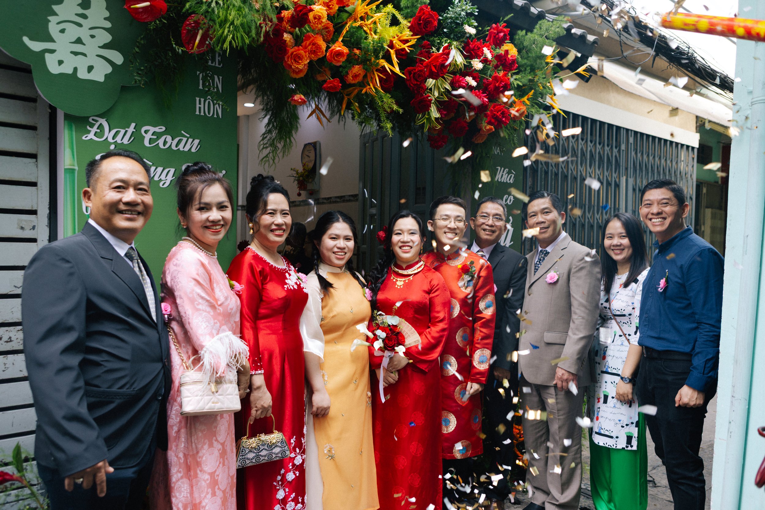 A group of people celebrating a wedding outside a building, with floral decorations and confetti in the air, posing for a photo.