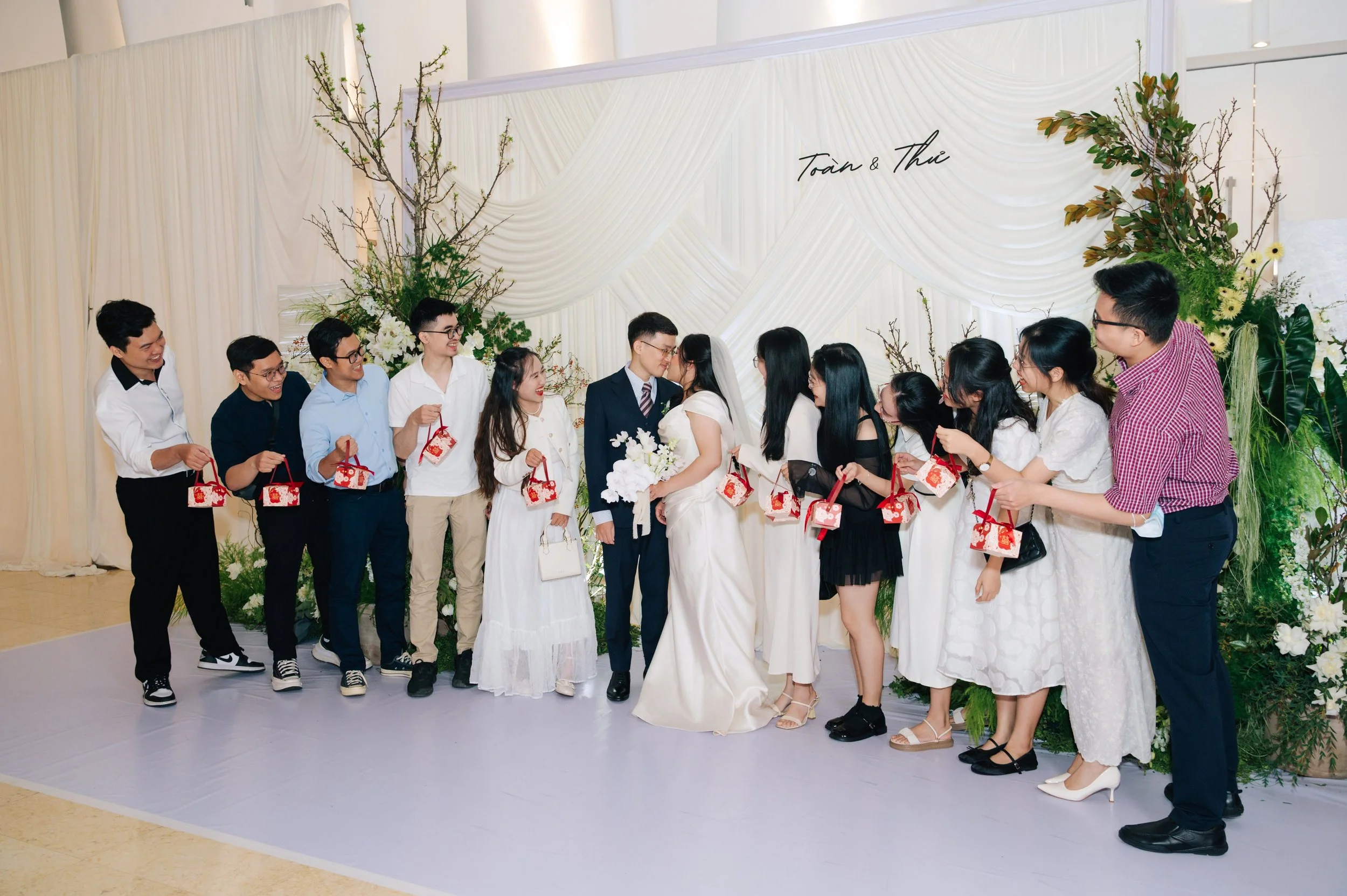 A wedding ceremony with the bride and groom kissing under a floral arch, surrounded by friends in a semi-circle holding small gift bags.