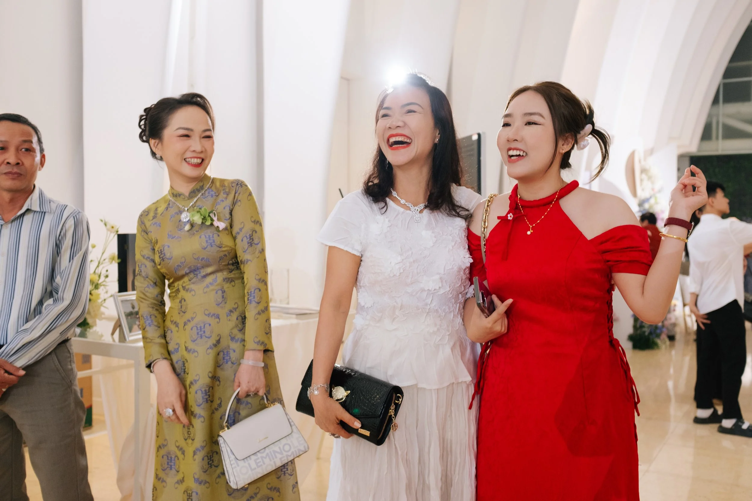 Four people standing together at an indoor event, smiling and enjoying the moment. The woman in the white dress and the woman in the red dress are sharing a laugh, while another woman in a yellow dress and a man in a striped shirt are also present.