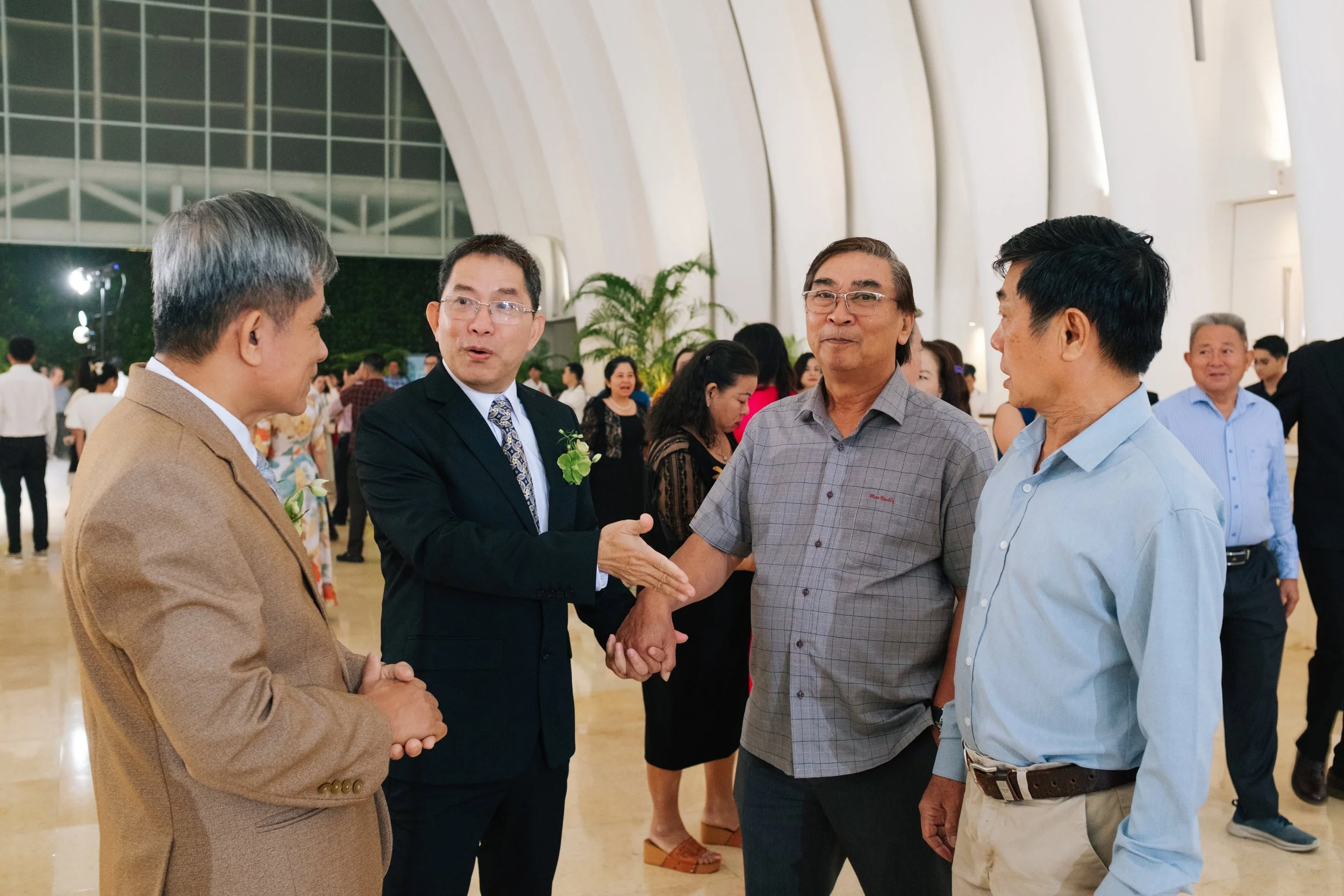 Four men engaged in conversation at an indoor event, with one man shaking hands with another, all dressed in semi-formal attire, surrounded by other people in the background.