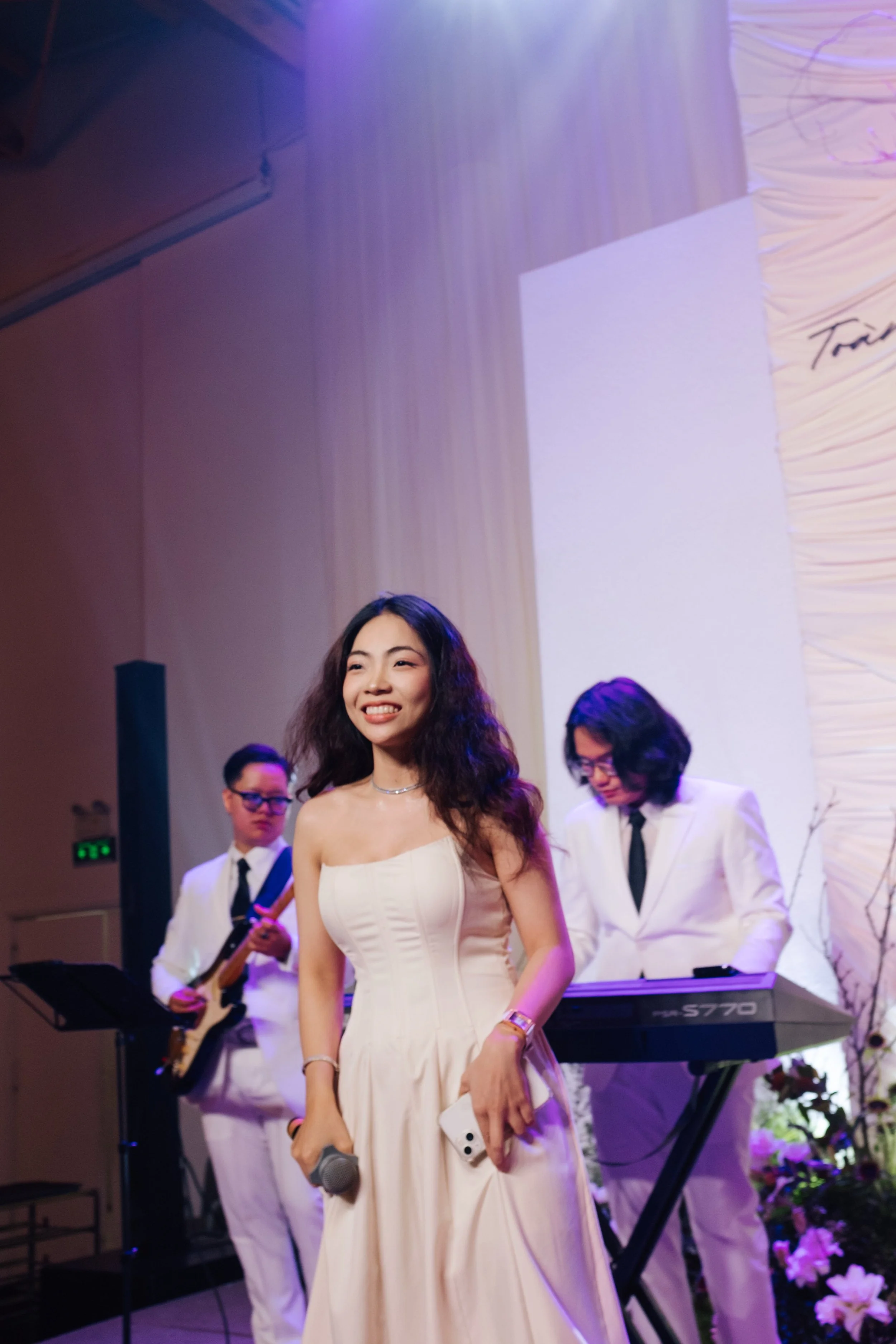A woman in a white dress smiling on stage with two men in white suits playing musical instruments behind her.