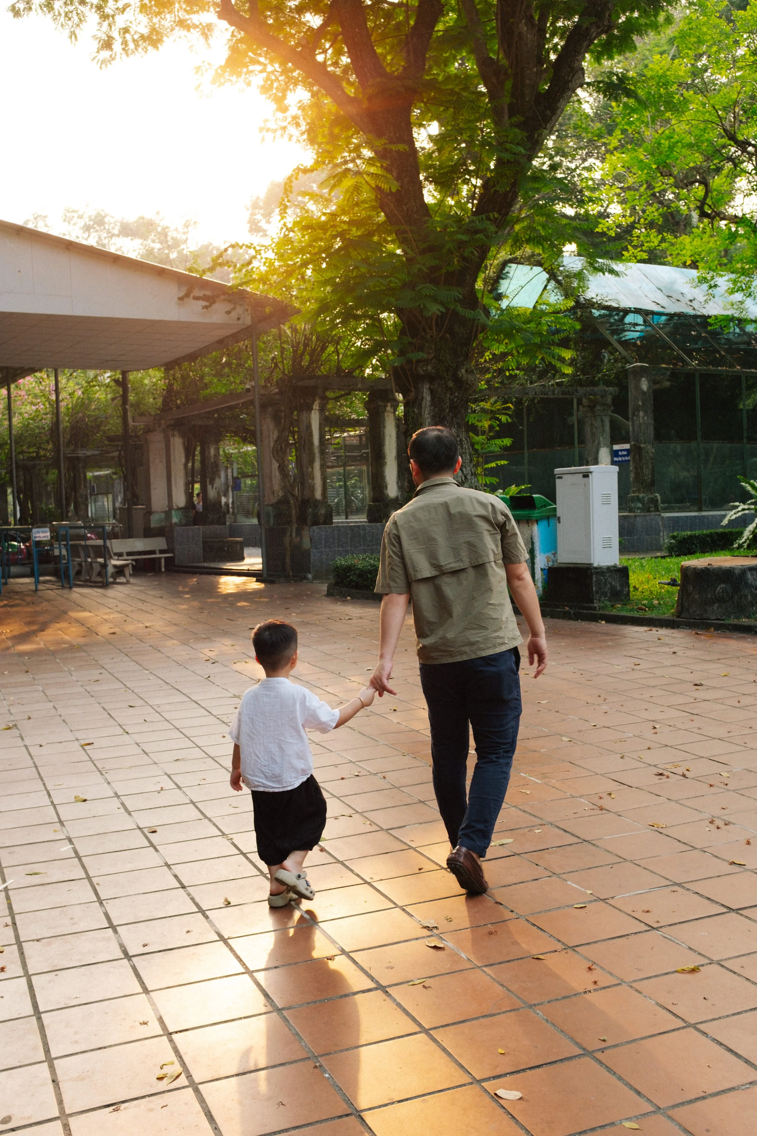 A man and a young boy walking hand in hand on a tiled path in a park during sunset, surrounded by green trees and park structures.