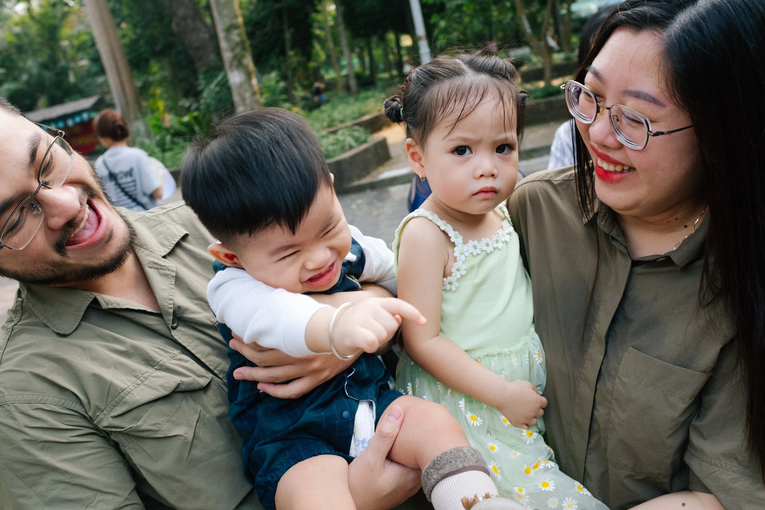 A family of four smiling and laughing outdoors in a park. The father is holding a laughing boy, the mother is smiling at a girl with serious expression, and all are dressed casually.