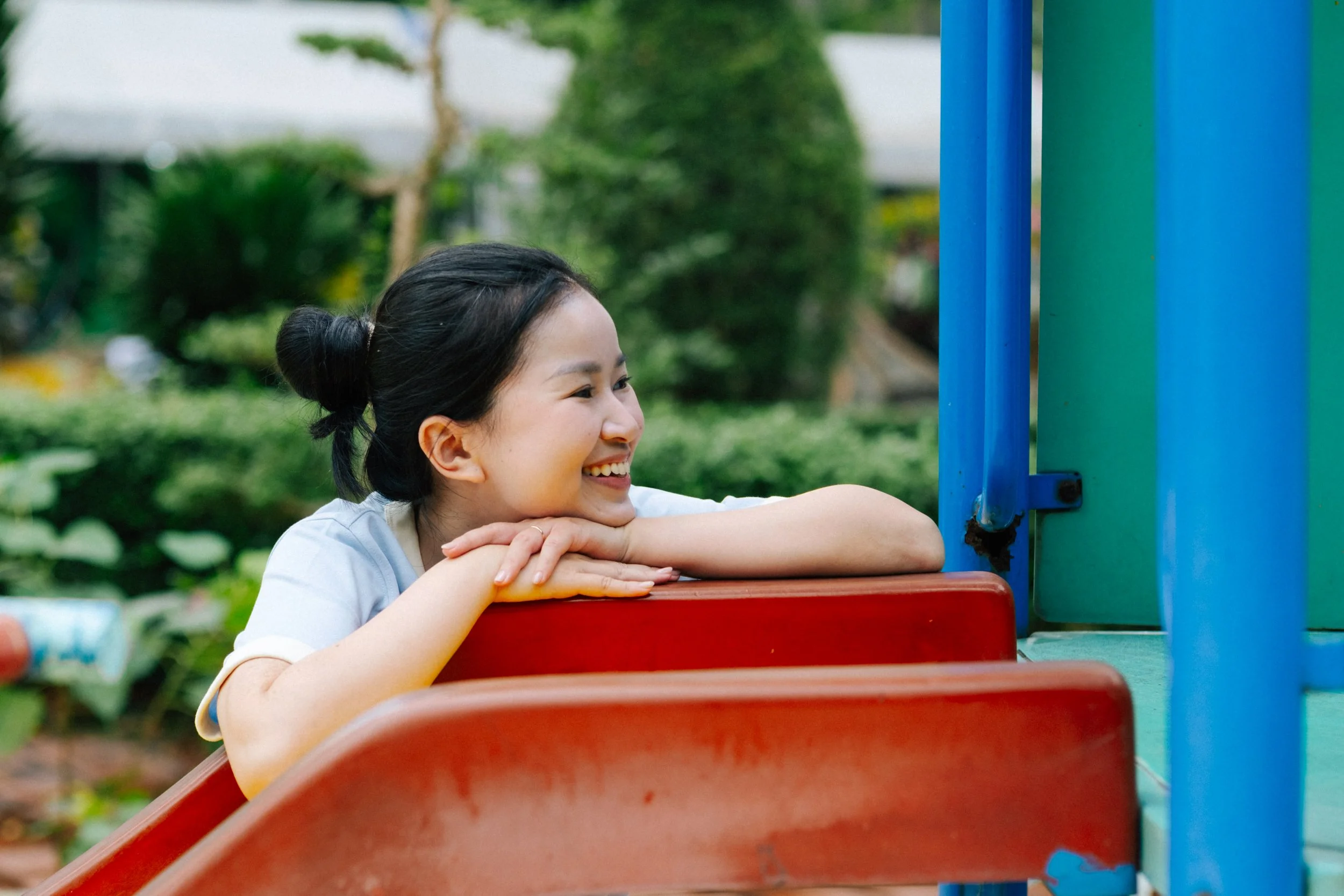 Young woman with dark hair in a bun smiling while leaning on playground equipment in a park.