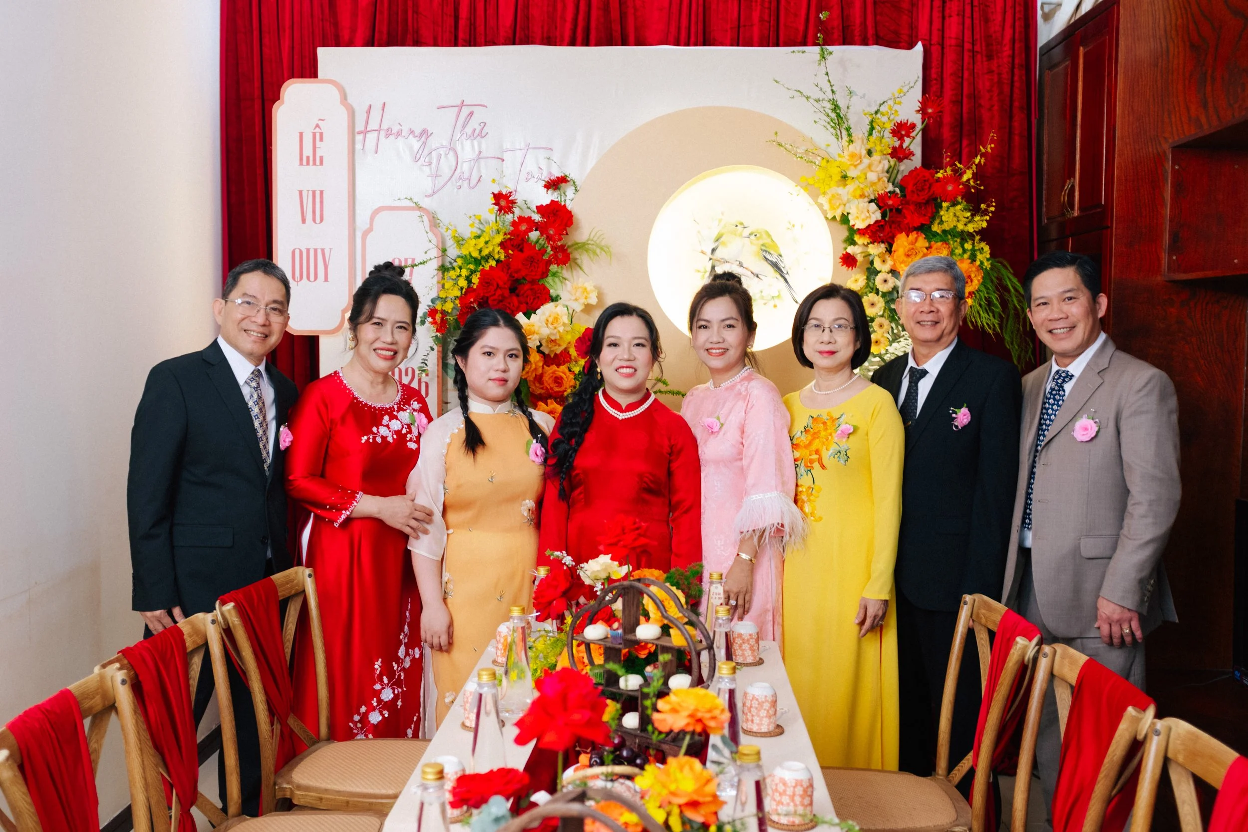 A group of nine people, including men and women, dressed in colorful traditional Vietnamese attire and formal suits, standing around a decorated table with flowers and food at a celebration.
Background features a red curtain, a white backdrop with ca
