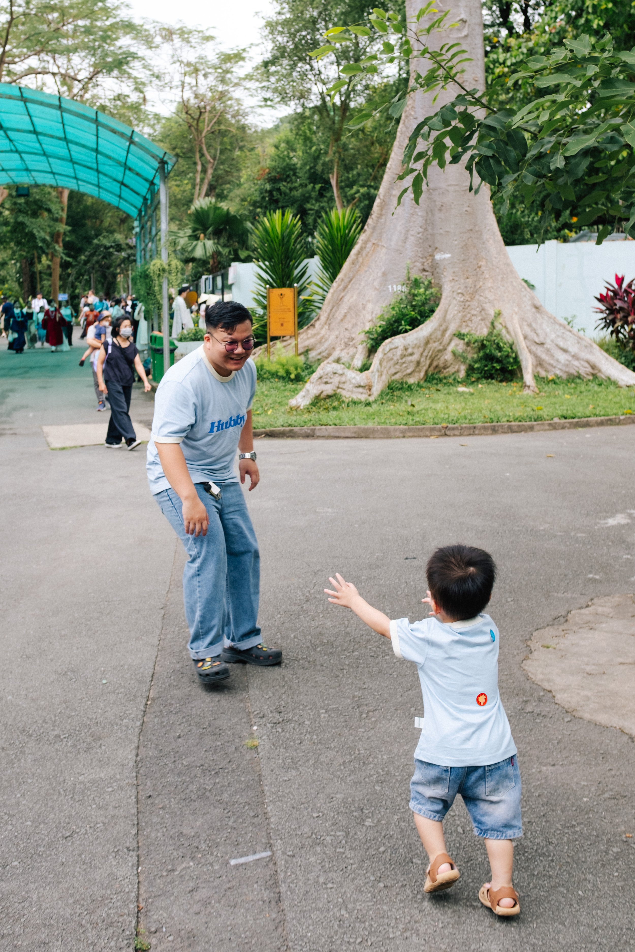 A man and a young boy playing at a park with a large tree and other visitors in the background.