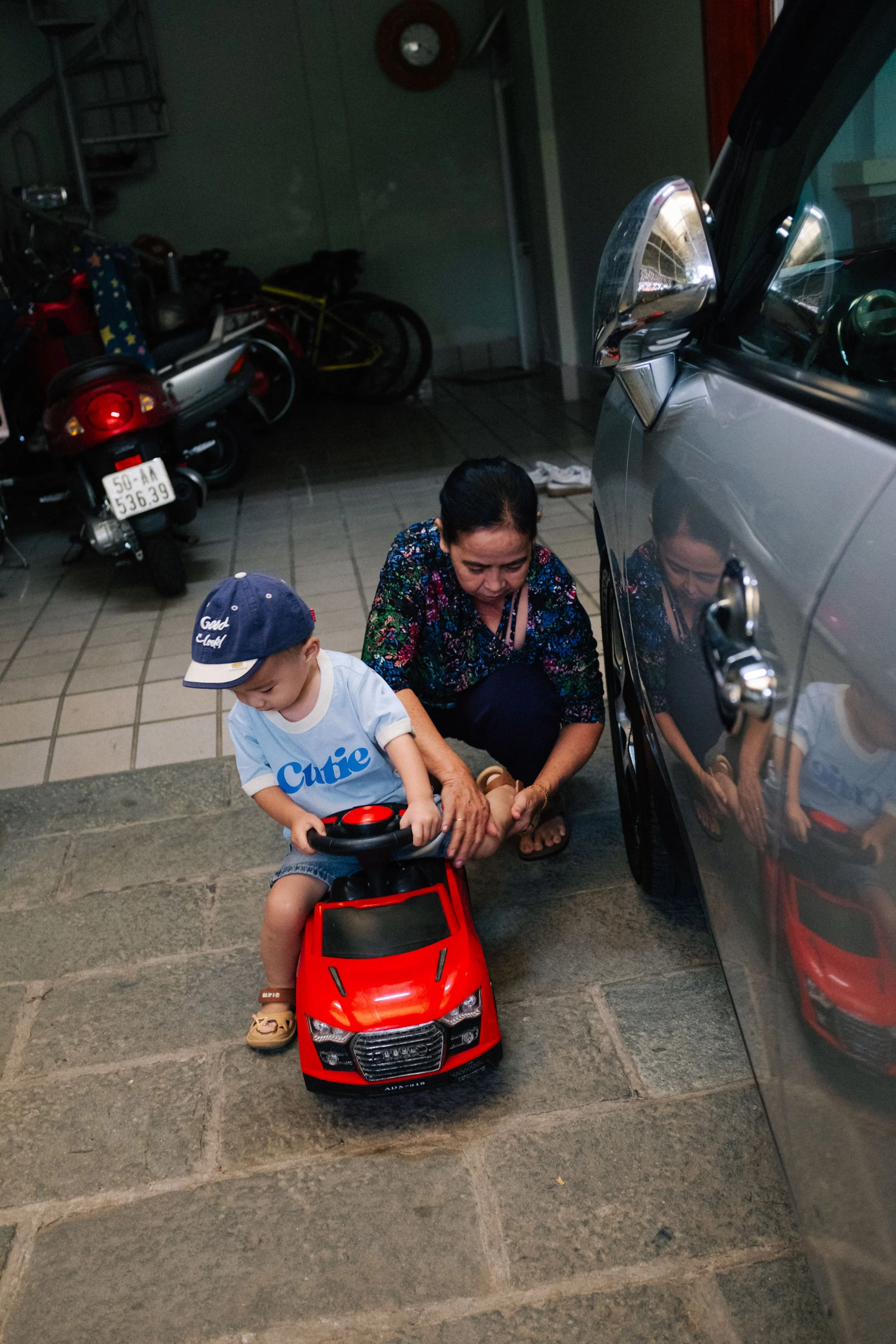 A young boy wearing a blue cap and a white shirt with the word 'Cute' on it, sitting on a red toy car, while an older woman crouches beside him, holding his leg, in a garage. There are motorcycles, a bicycle, and a gray car with its side mirror visib