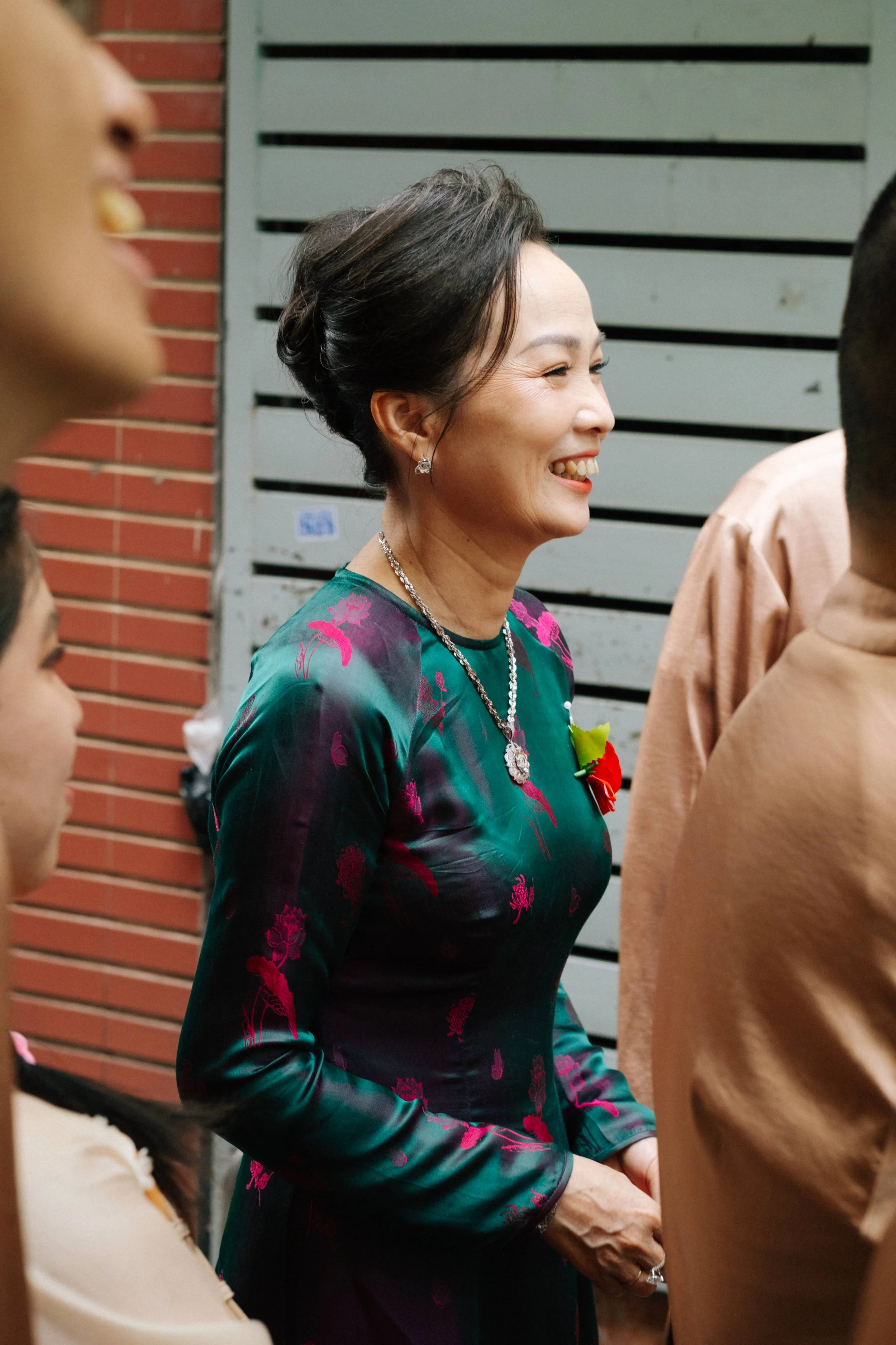 A smiling woman dressed in a traditional Vietnamese dress with floral patterns, wearing jewelry and a boutonniere, standing among a group of people indoors or in a sheltered area.