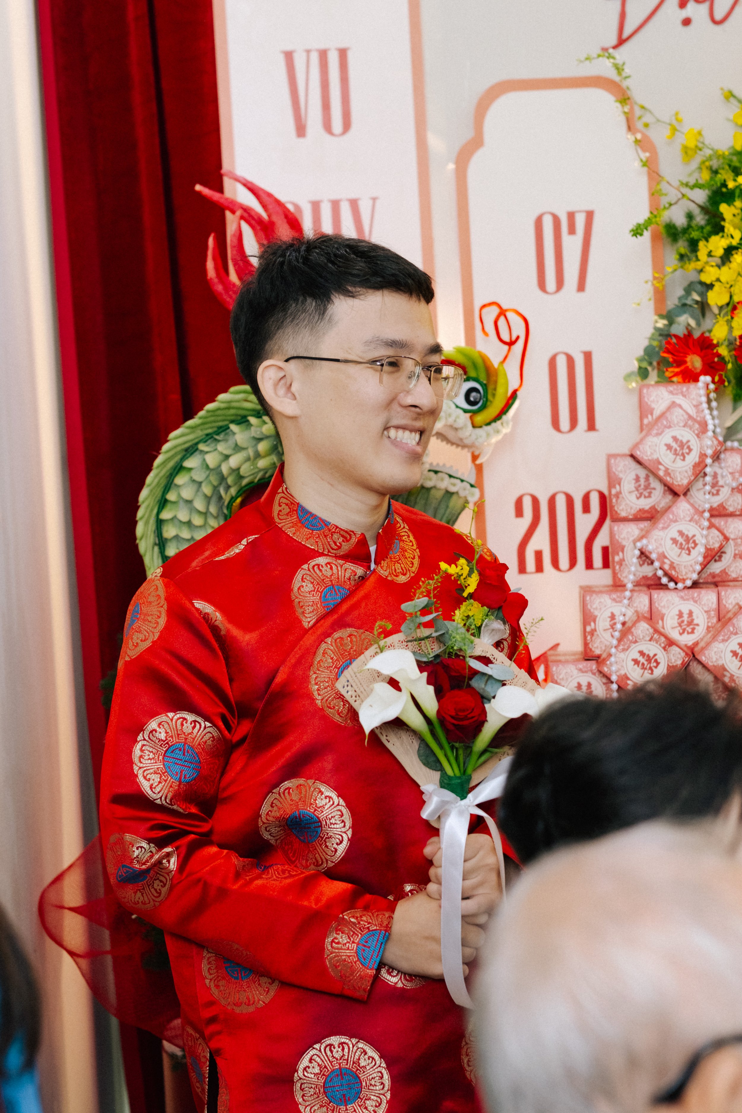 A young man wearing glasses and a red traditional Chinese outfit holds a bouquet of red and white flowers while smiling during a celebration. Behind him, there are Chinese decorative elements and red envelopes, indicating a festive event.