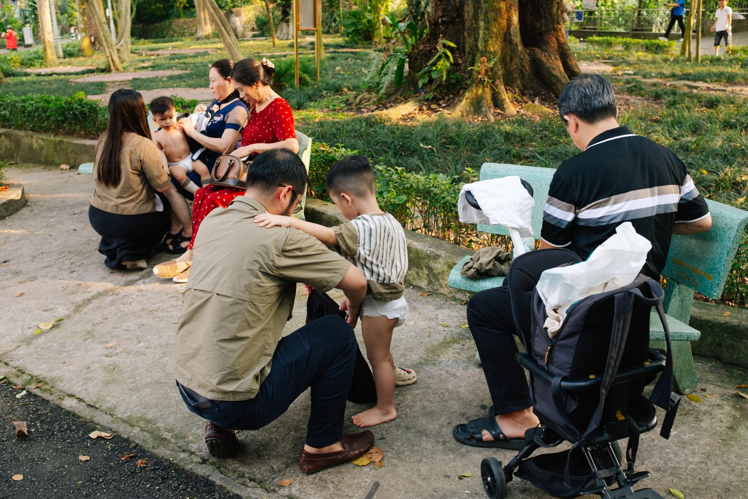People sitting on park benches and sitting on the ground in a park, some tending to children.