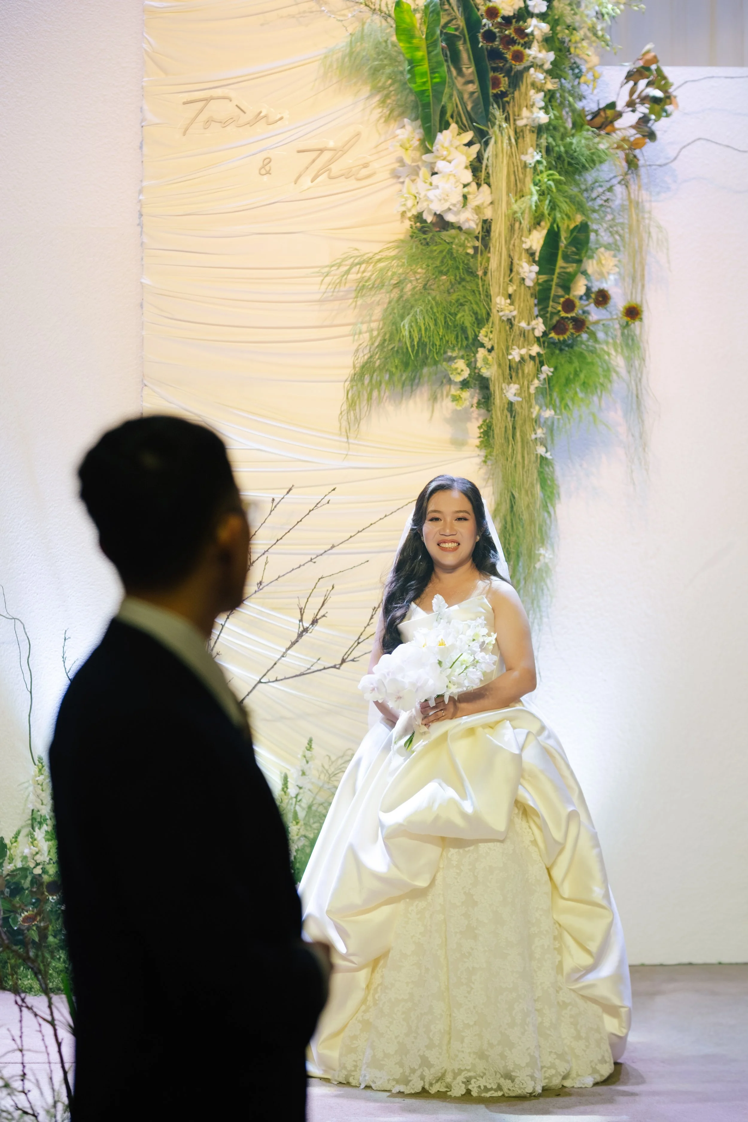 A bride in a white wedding gown holding a bouquet of white flowers, smiling and sitting on a pedestal at her wedding. A groom in a black suit stands in the foreground, blurred out, during their wedding ceremony with a floral backdrop.