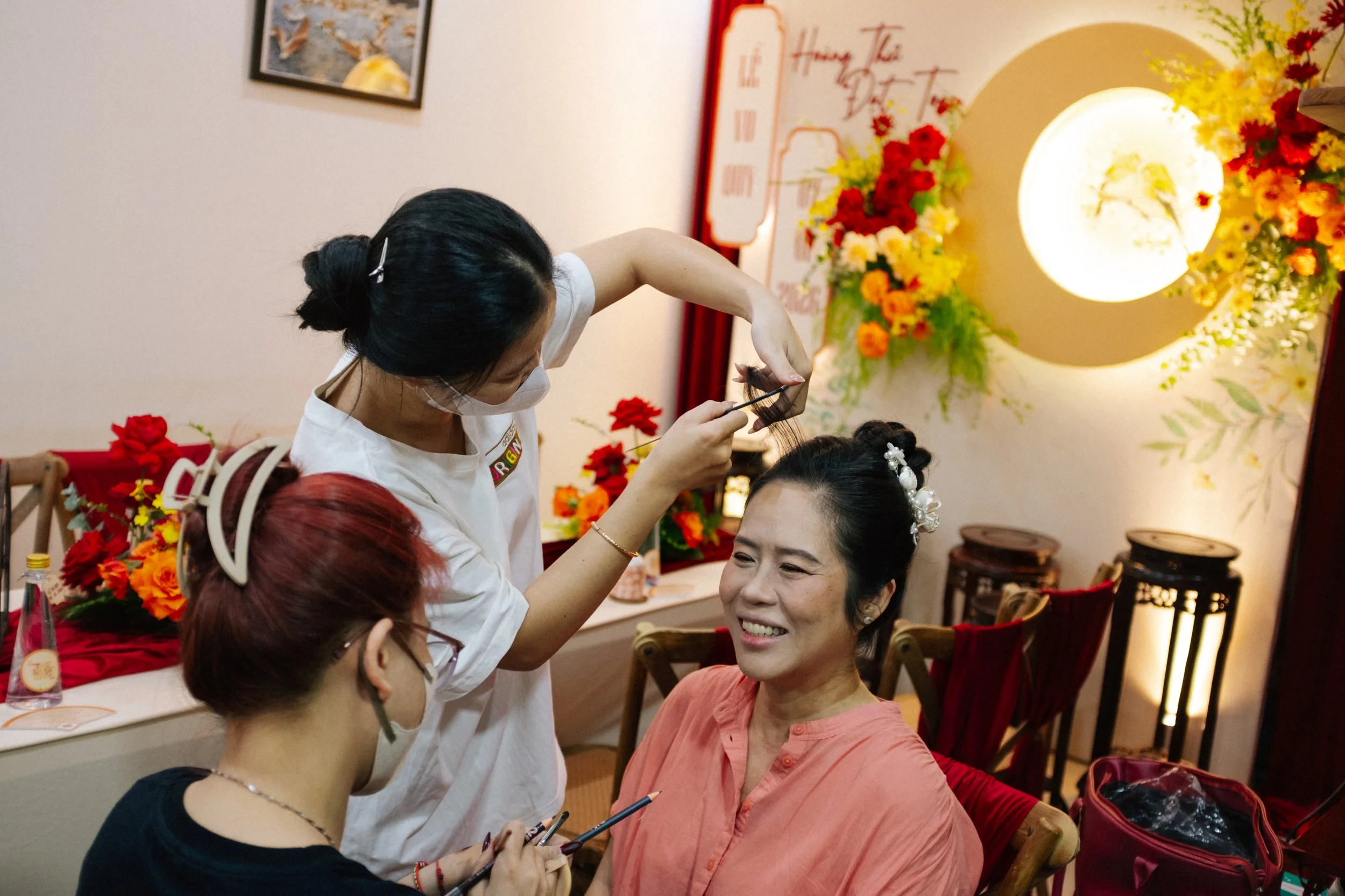 A woman getting her makeup done, seated in a chair, with two makeup artists working on her; one artist is applying makeup while the other is holding makeup tools. The scene is decorated with red and yellow flowers and warm lighting.