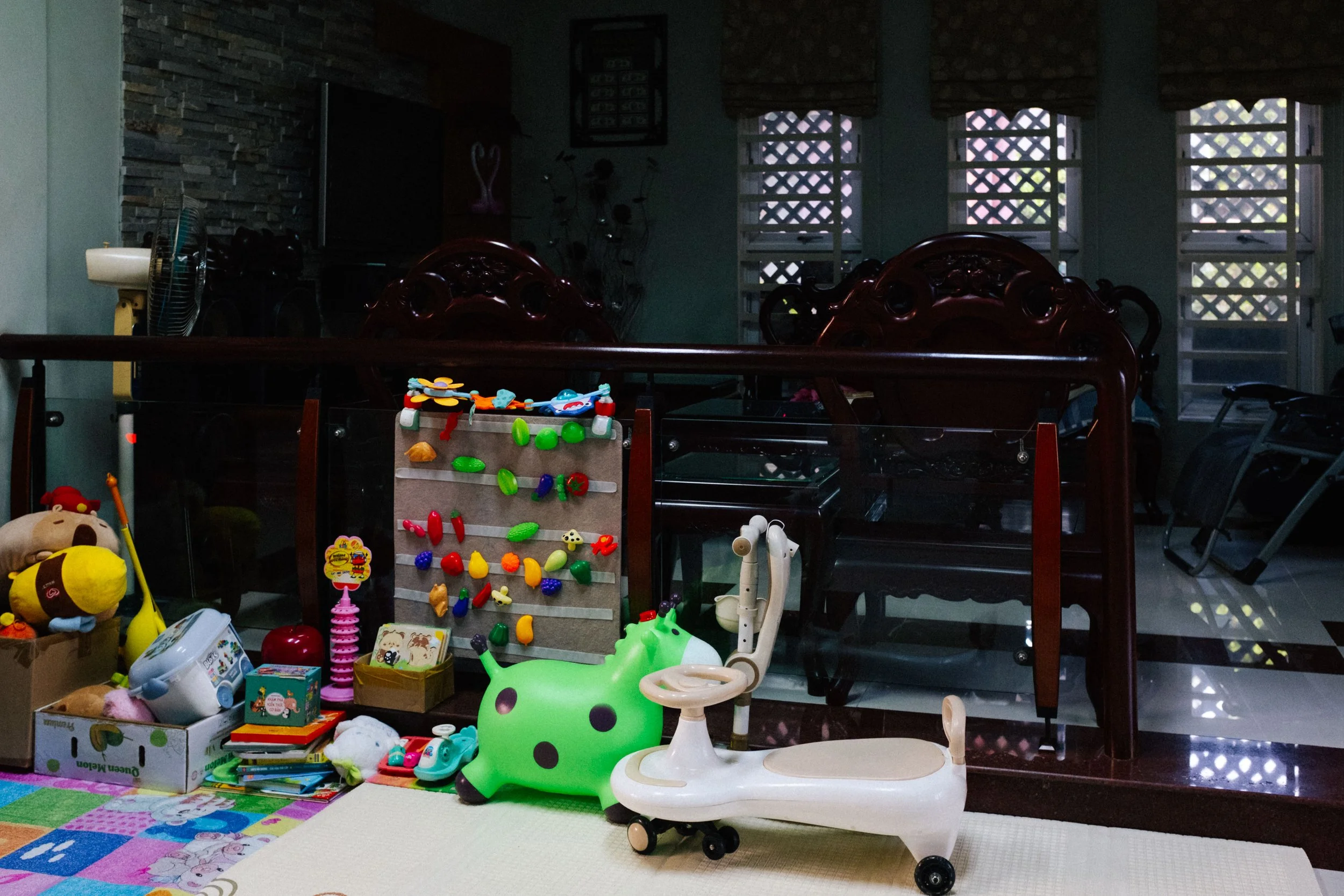 Child's play area with toys including a green inflatable toy, a wooden ride-on, and a colorful pegboard with miniature toys, in front of a dark wooden railing and a living room with windows, a TV, and various items in the background.