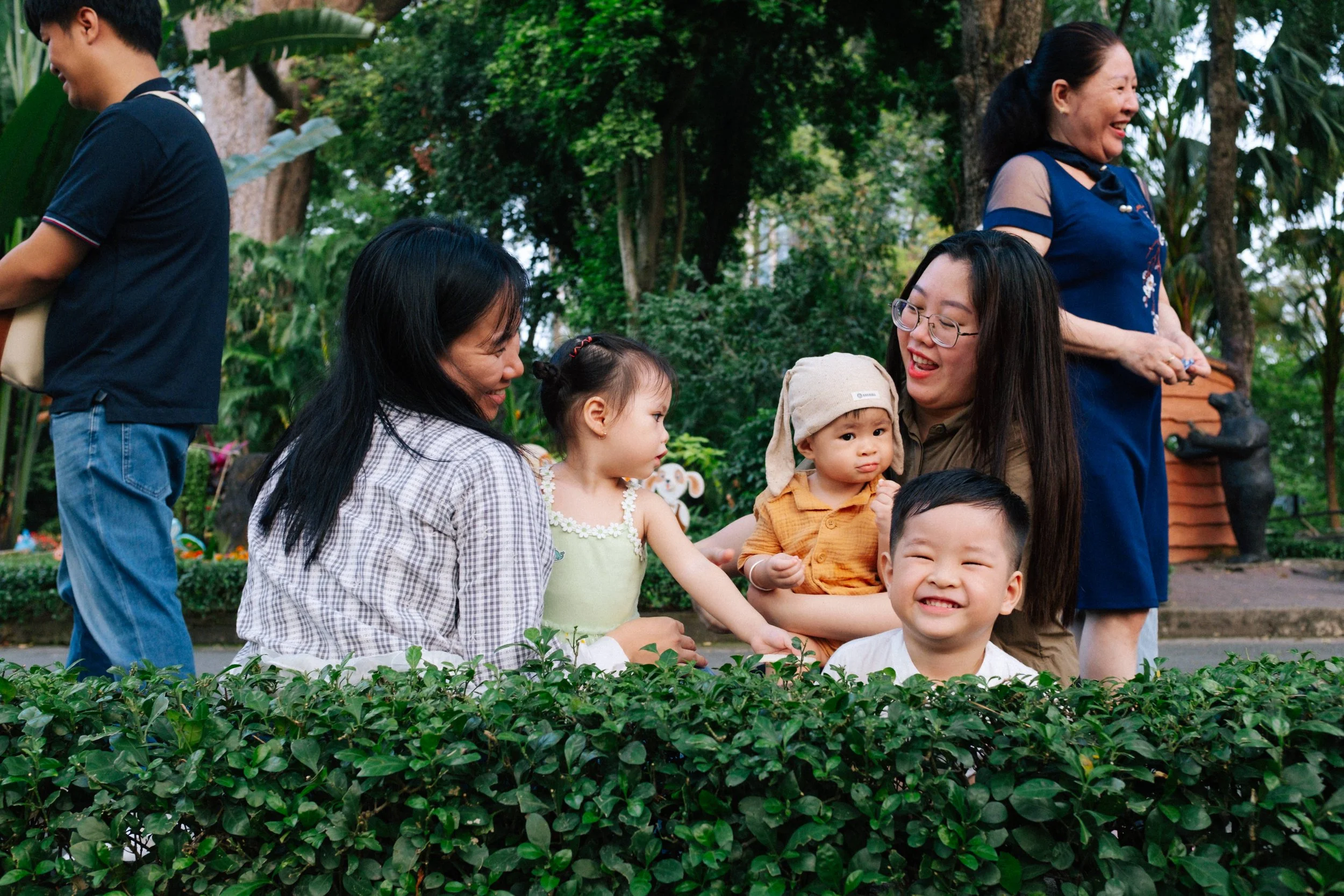 Group of children and women smiling and engaging in outdoor activities in a garden with lush greenery and trees.